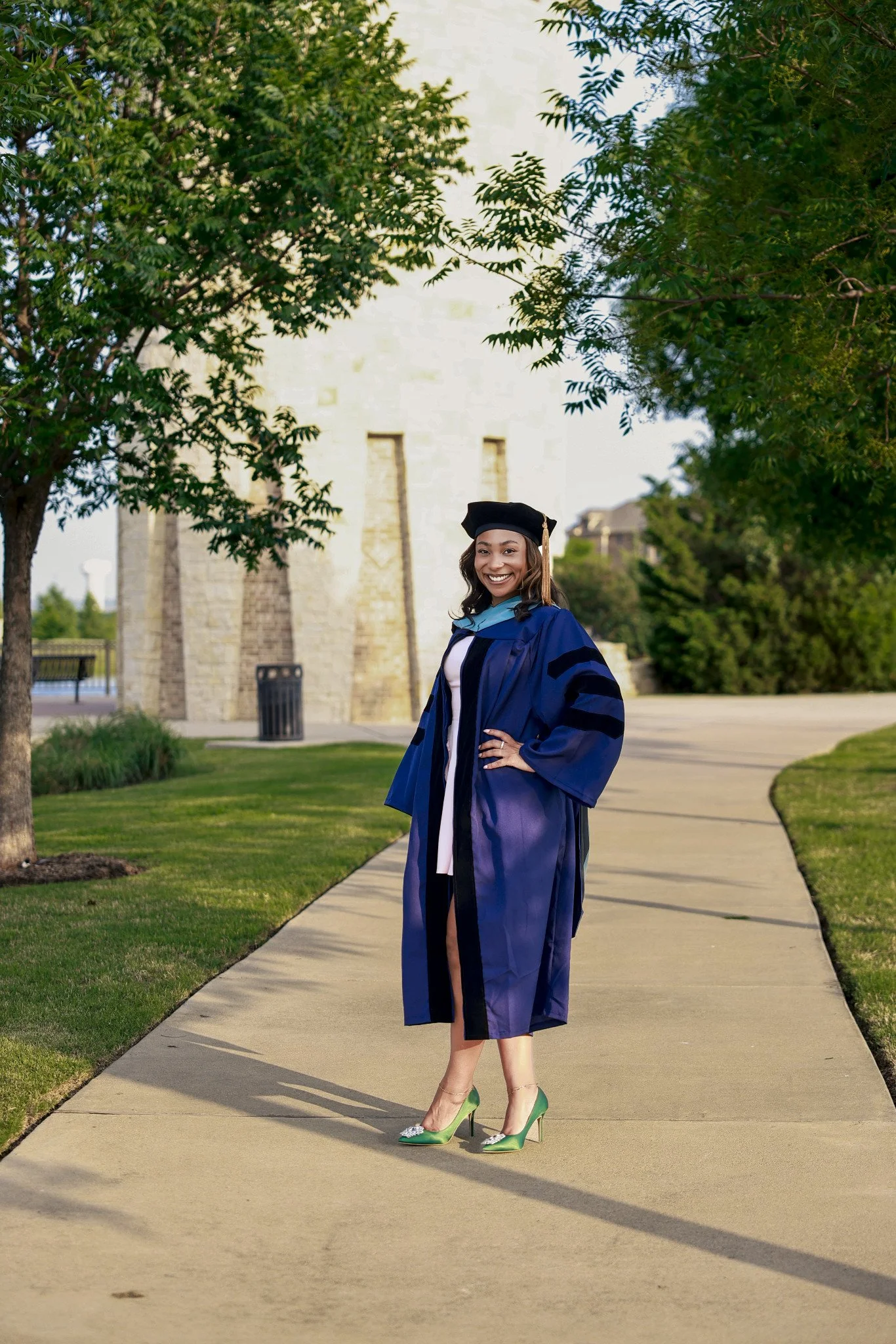 A woman standing outdoors on a sidewalk, wearing a blue graduation gown and cap, smiling, with greenery and a stone building in the background.
Milestone Image