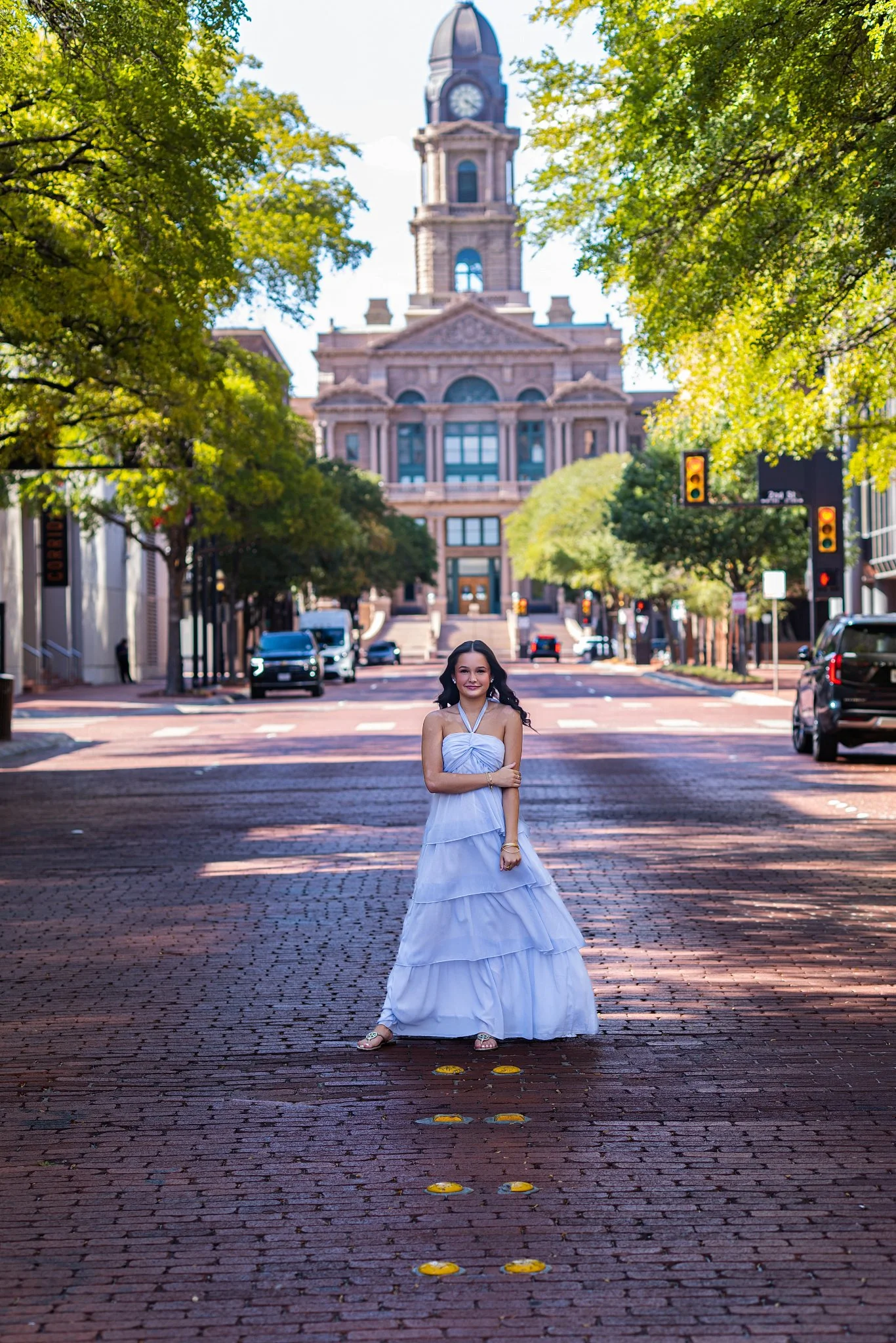 A woman in a long white dress stands on a brick street in a city, with a historic building with a clock tower in the background, surrounded by green trees and a traffic signal.