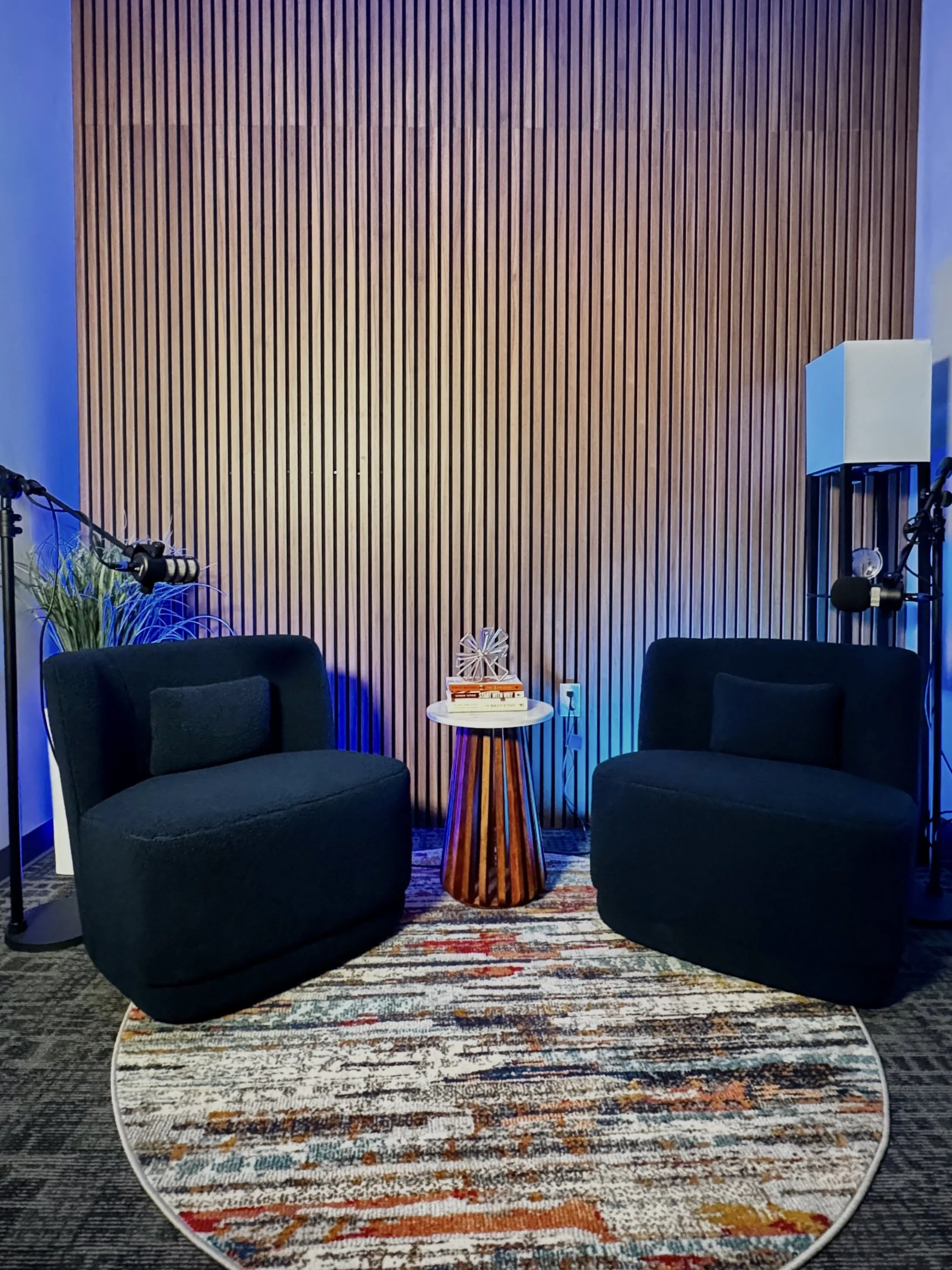 A studio setup with two black upholstered chairs facing each other on a colorful circular rug, a small wooden side table with books and a decorative sculpture between them, and soundproofing panels on the wall. Microphones and studio equipment are visible on the sides.