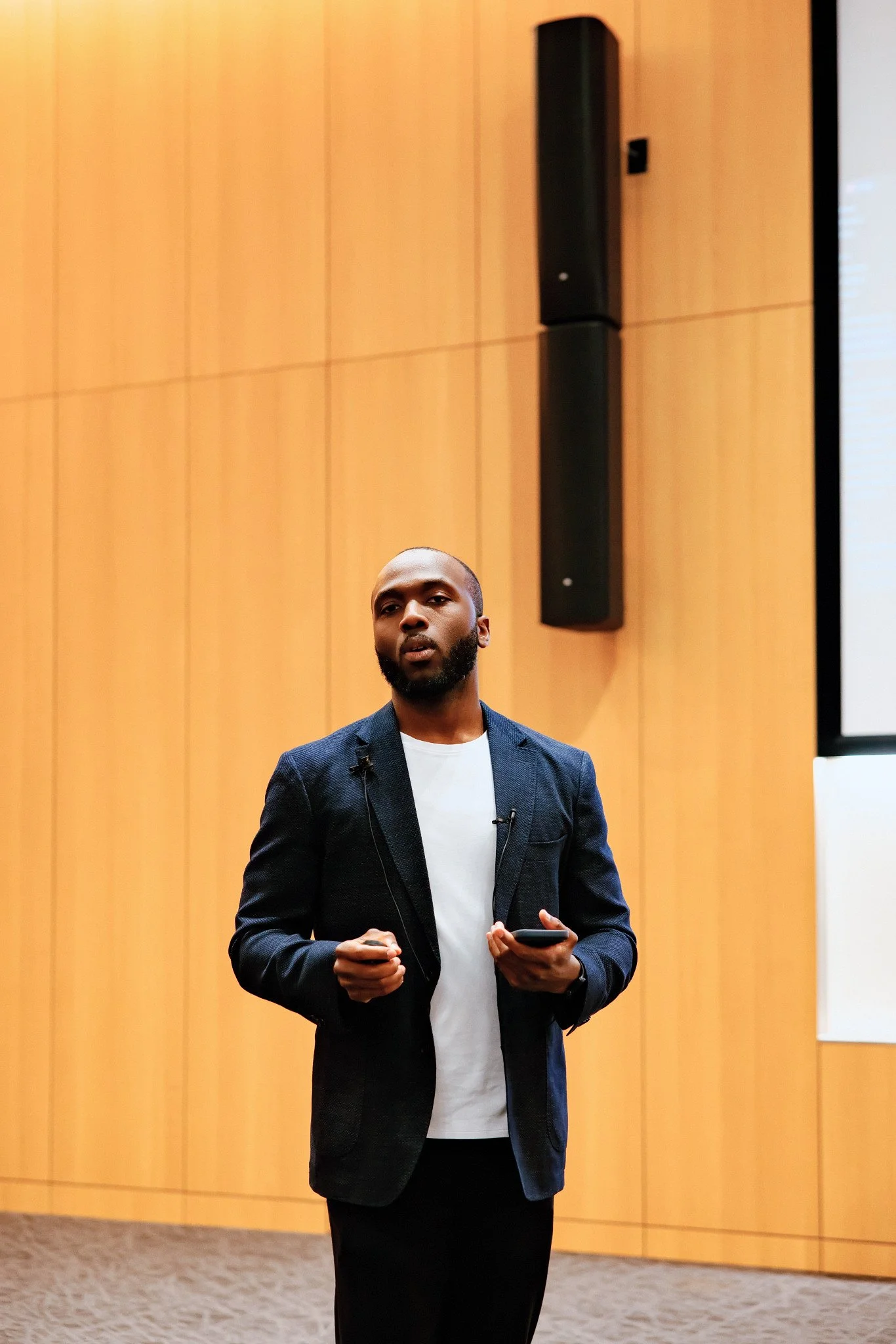 A man with a dark beard and wearing a dark blazer over a white shirt stands on a stage or in a conference room with wooden paneling. He appears to be speaking or giving a presentation, holding a phone in his right hand.