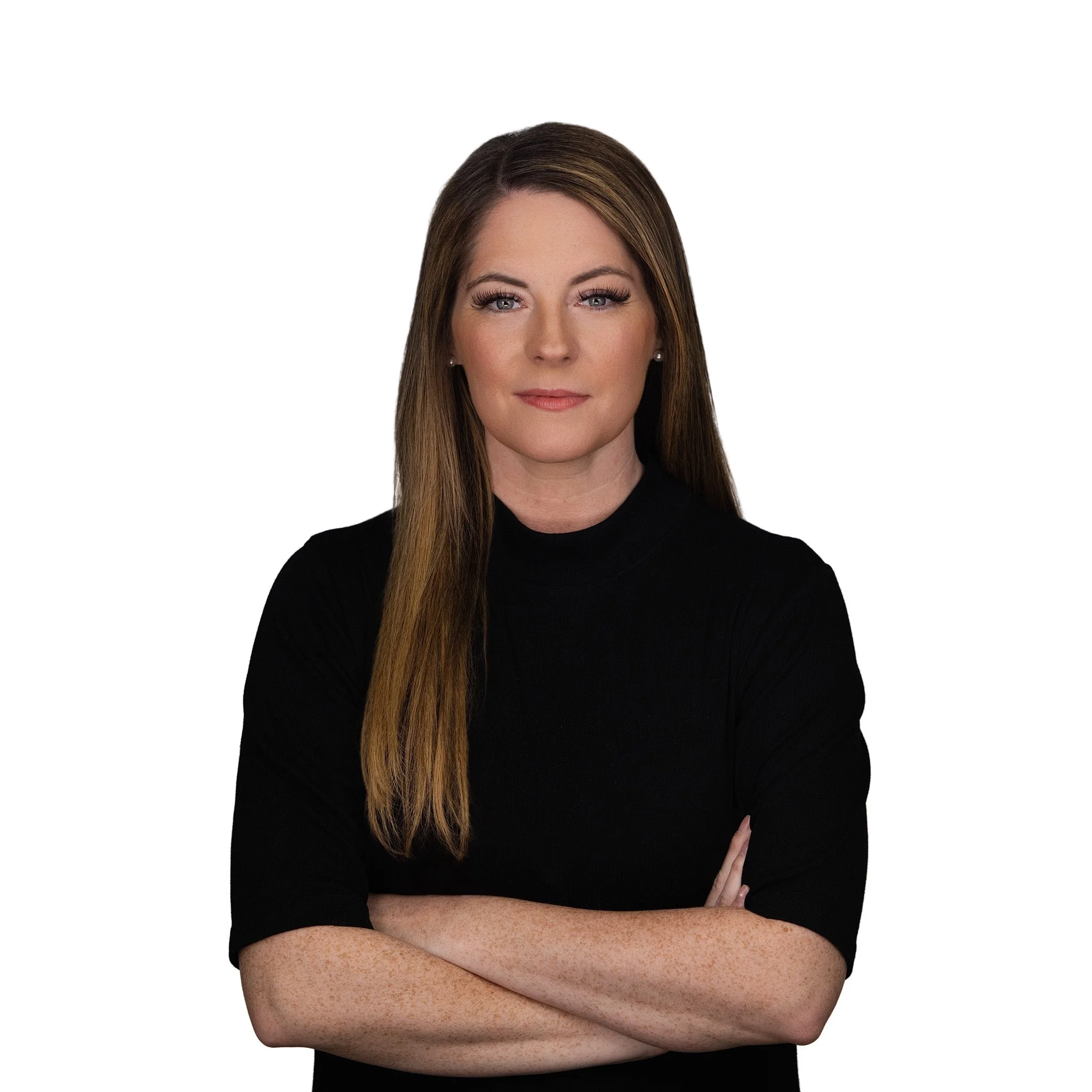 A woman with long brown hair, wearing a black top, stands with her arms crossed against a plain white background. Executive Headshot