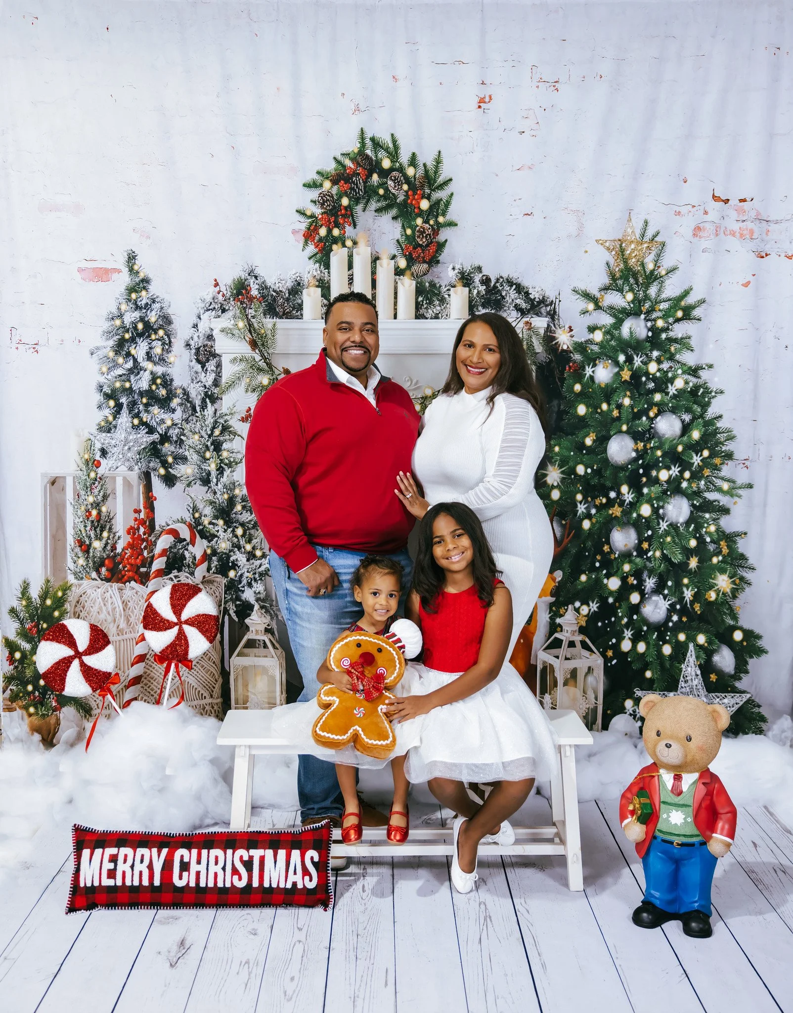 A family celebrating Christmas indoors with decorated Christmas trees, holiday ornaments, and festive props. A man in a red sweater and a woman in a white dress stand behind two children, one in a red dress. The children hold Christmas-themed plush toys and gingerbread decoration. There are wrapped presents, lanterns, and Christmas decorations on the floor, with a large "Merry Christmas" sign.