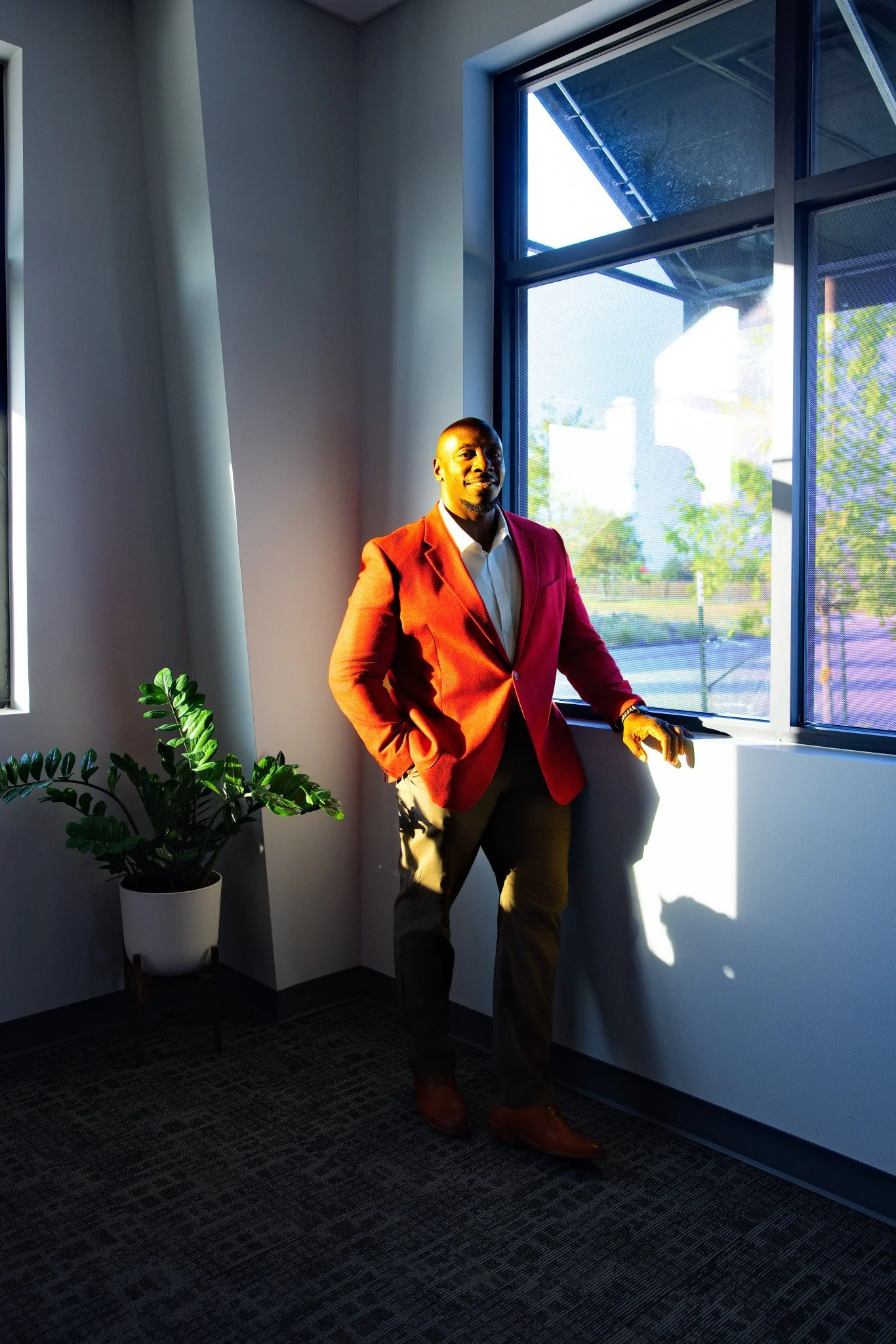 Man in a red blazer standing by a large window with sunlight streaming in, next to a potted green plant, in an office space.