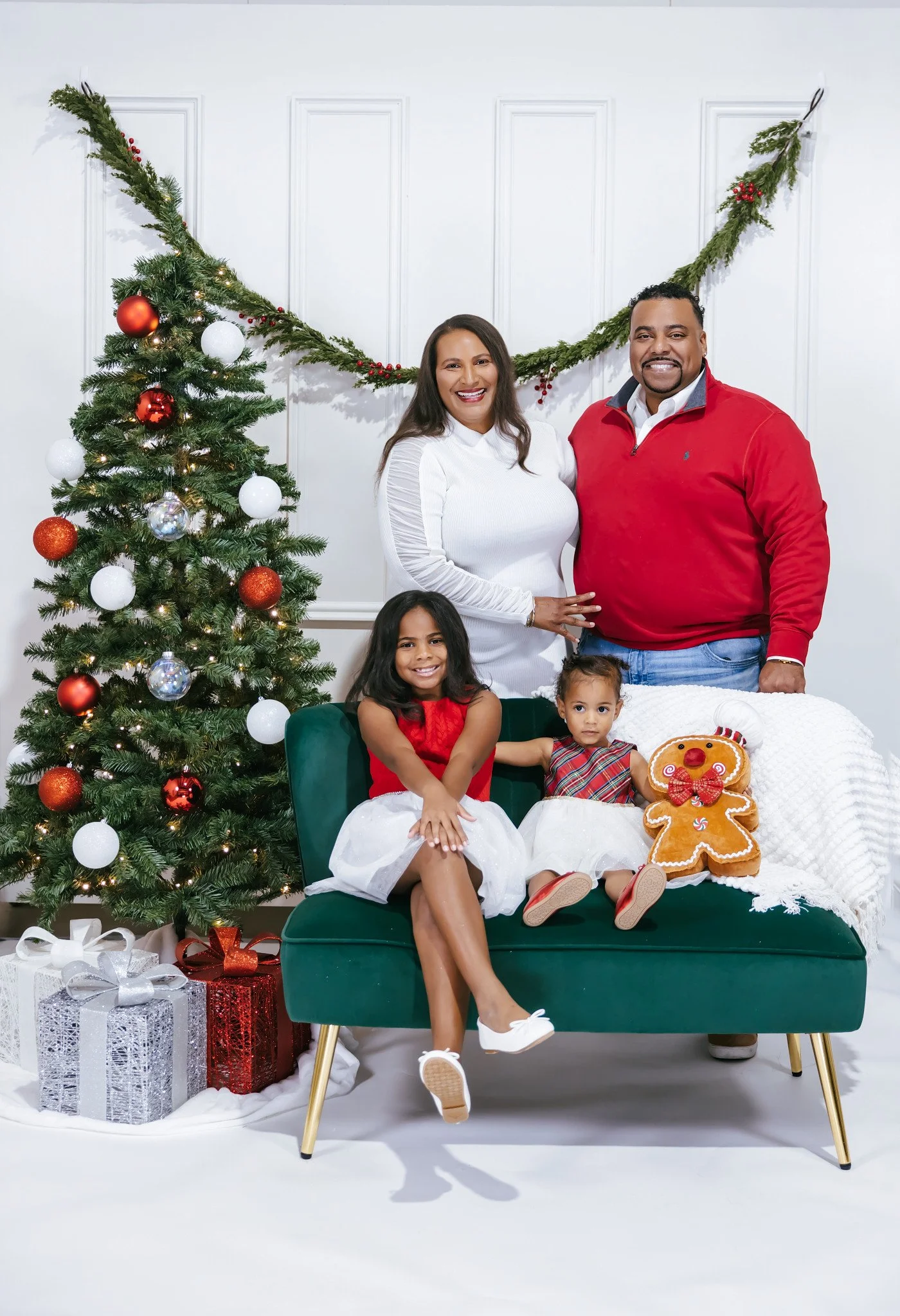 Family posing for Christmas photo with decorated tree, presents, and plush gingerbread man, in a white room.