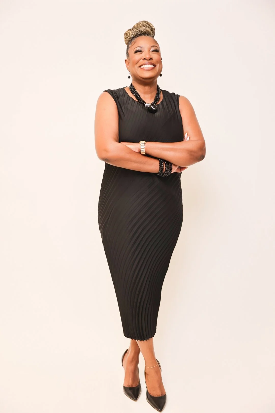 A woman standing with arms crossed, wearing a black pleated dress, black heels, and jewelry, smiling against a plain white background.