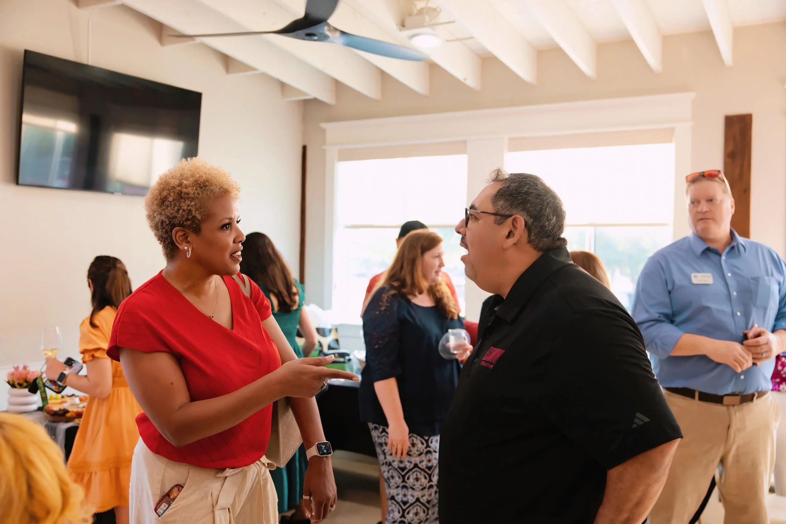 Two people engaged in a conversation at a social gathering, with others in the background, in a well-lit room.