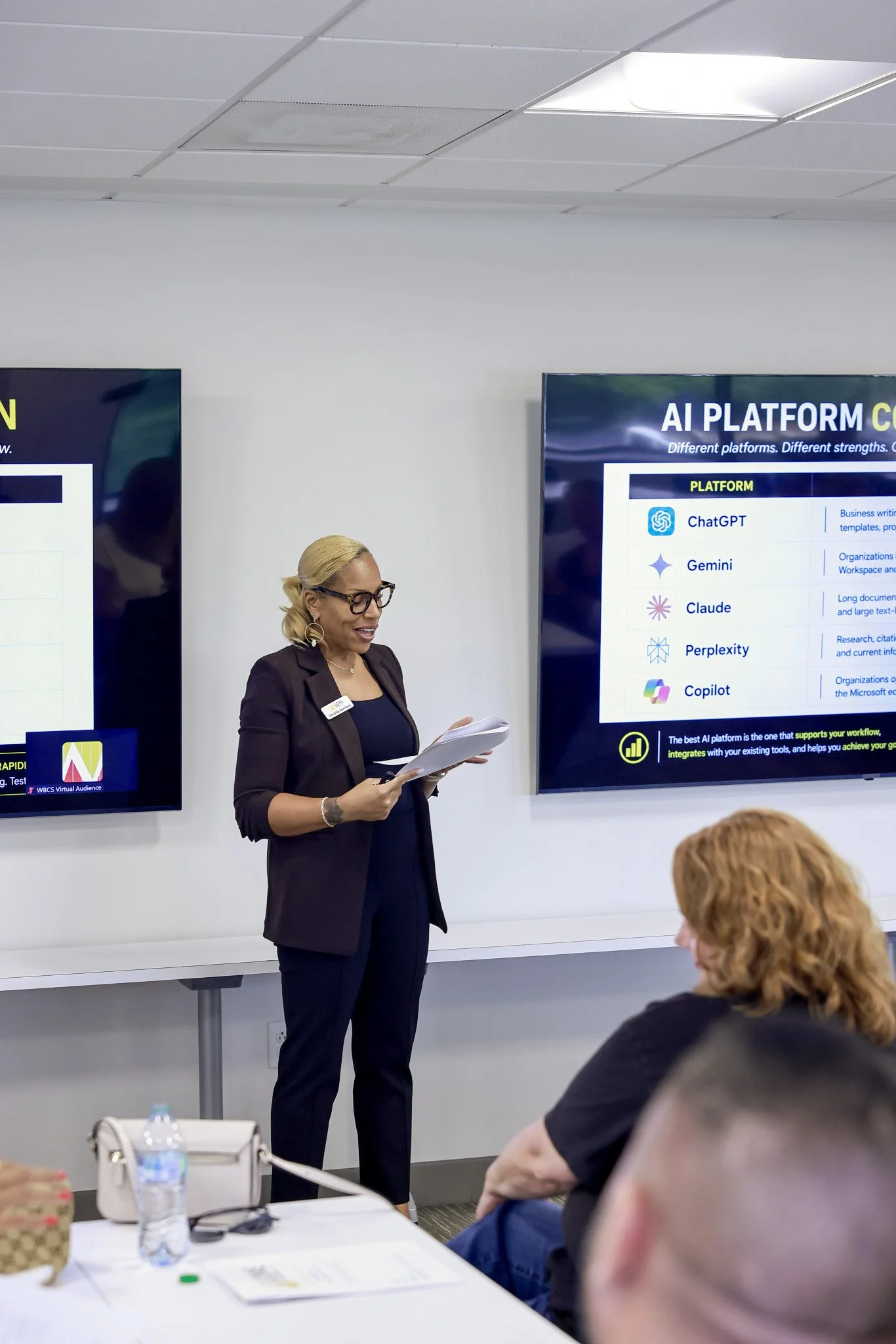 A woman with blonde hair, glasses, and earrings, standing in front of a large screen and giving a presentation to an audience. The screen displays a slide titled 'AI PLATFORM' with lists of platforms like ChatGPT, Gemini, Claude, Perplexity, and Copilot.