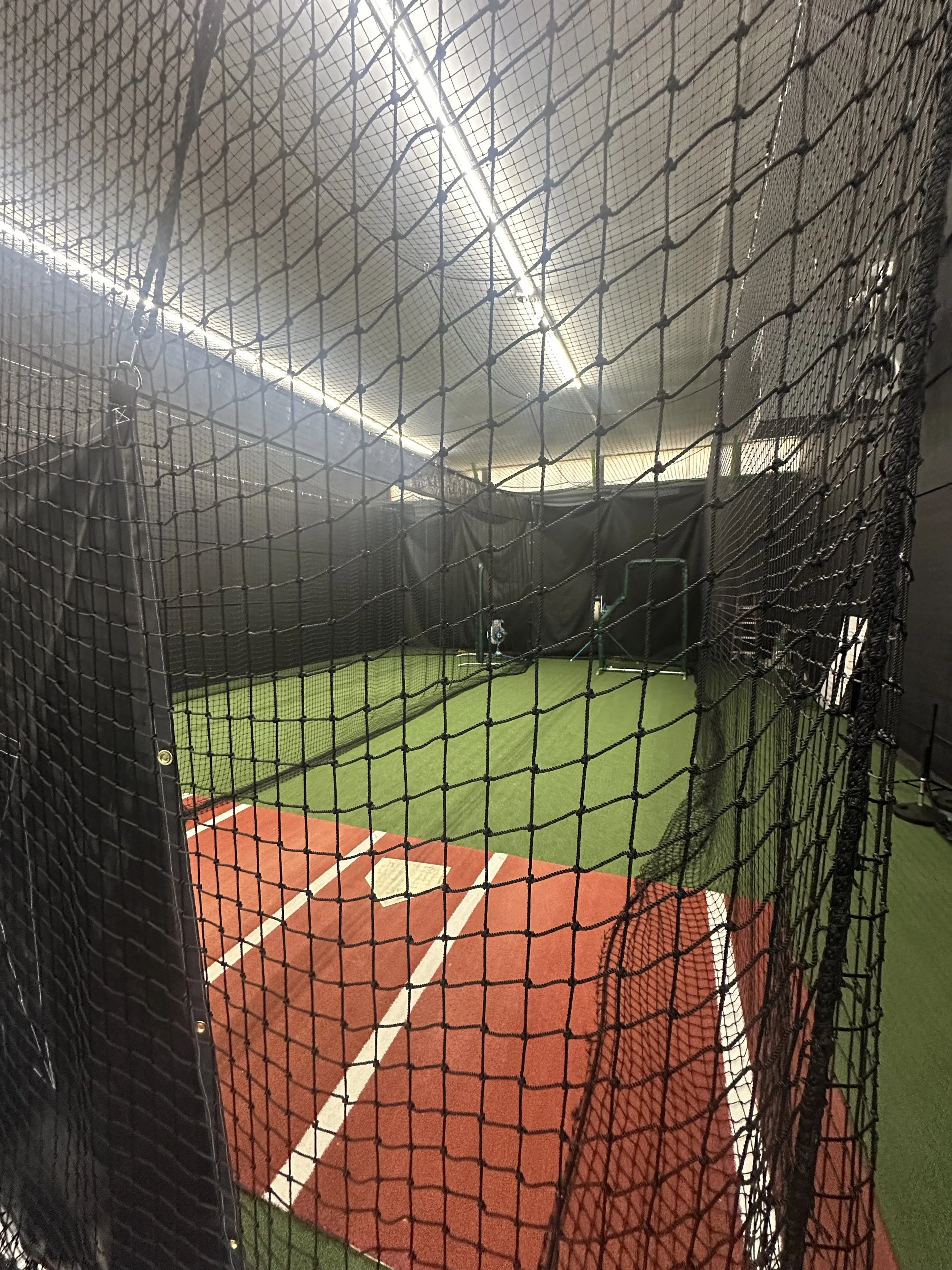 View of an indoor batting cage with a green artificial turf, a section of the red clay baseball/softball field, and netting surrounding the area.