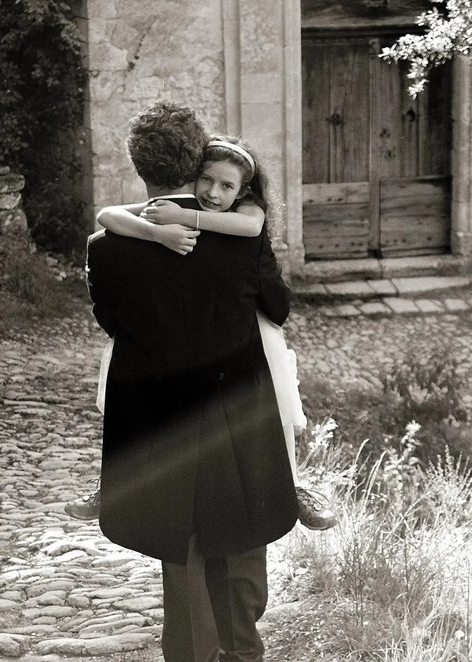 A man holding a young girl in his arms outdoors on a cobblestone path near a stone wall and wooden gate, with trees and plants nearby.