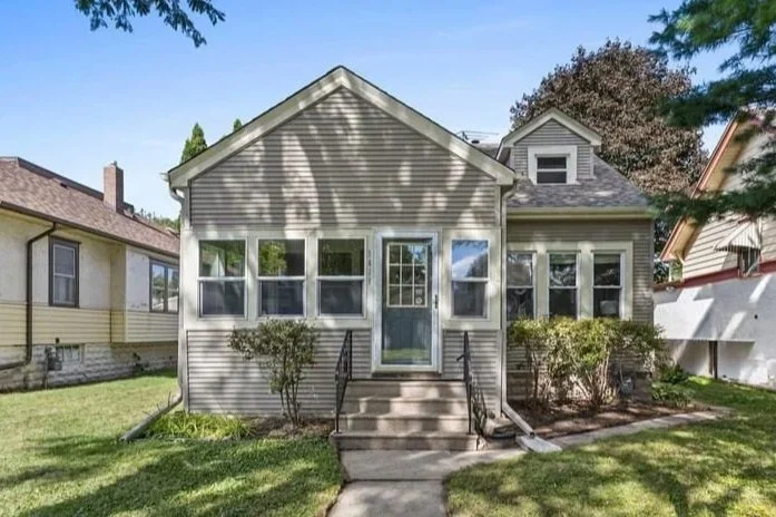 A two-story gray house with a screened-in front porch lit from inside, surrounded by trees with autumn-colored leaves at sunset. Recently sold Lowry Hill home.