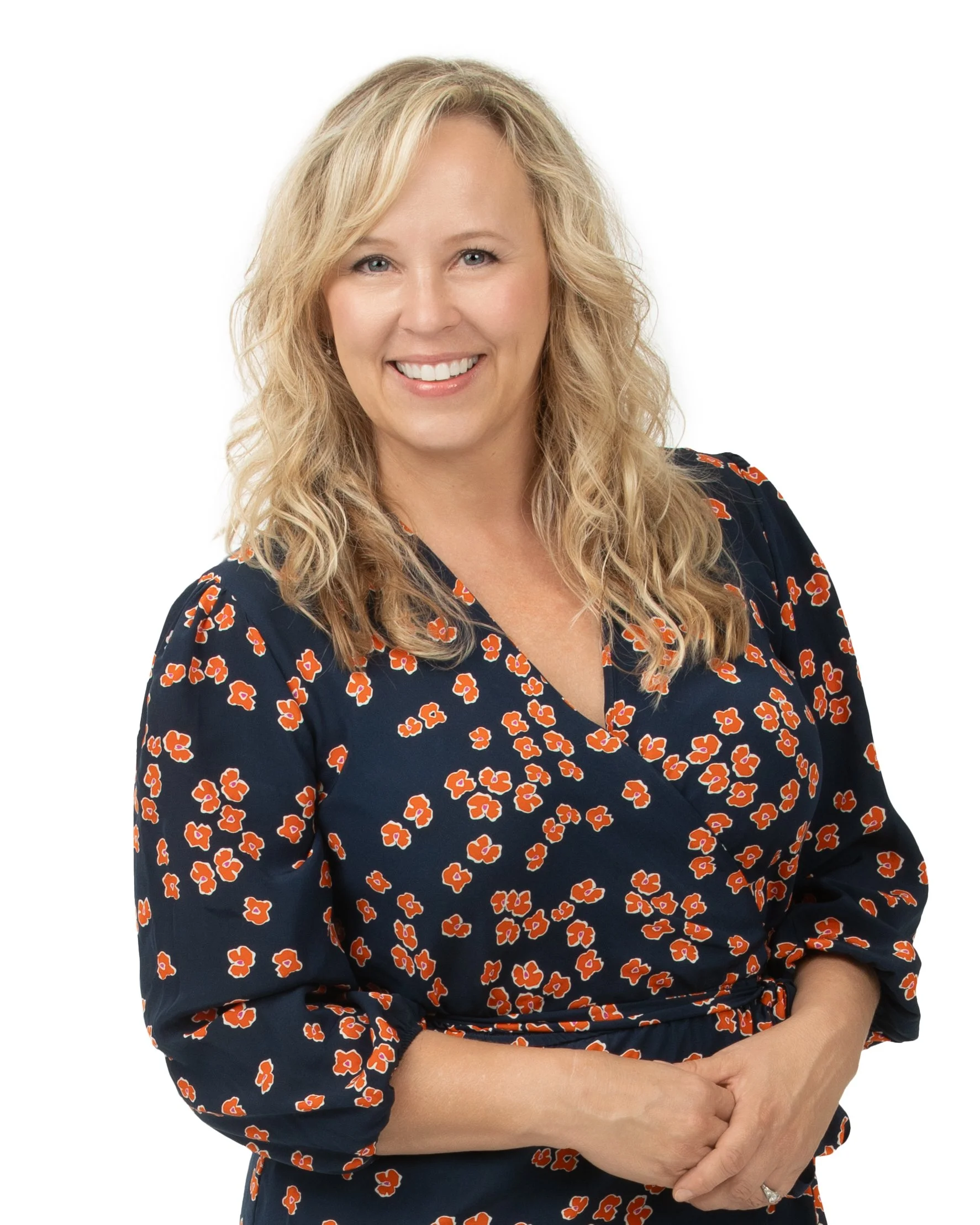 A smiling woman with blonde, wavy hair wearing a dark blue dress with a red floral pattern, standing against a white background. Woman is Jennifer Plourde Partner and Co-Founder of the Plourde Team along with Shannon and Maia. Director of operations.