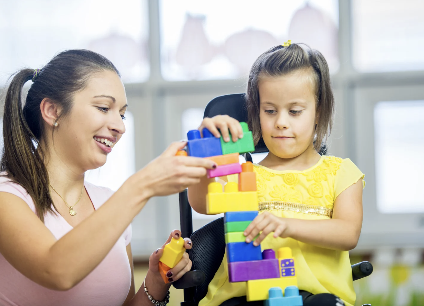 A young girl in a wheelchair stacking colorful toy blocks with a woman smiling and assisting her.
