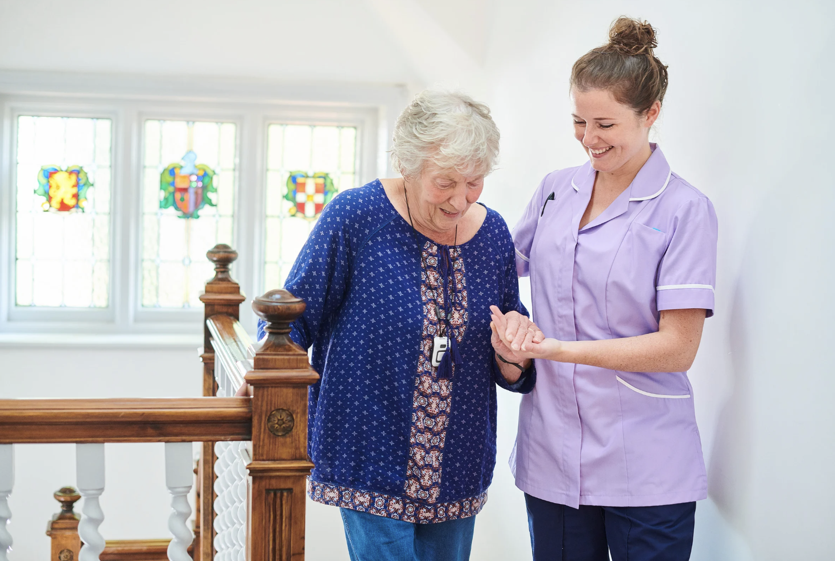 A young female healthcare worker in a purple uniform helping an elderly woman with a medical device on her wrist, standing on a staircase with stained glass windows in the background.