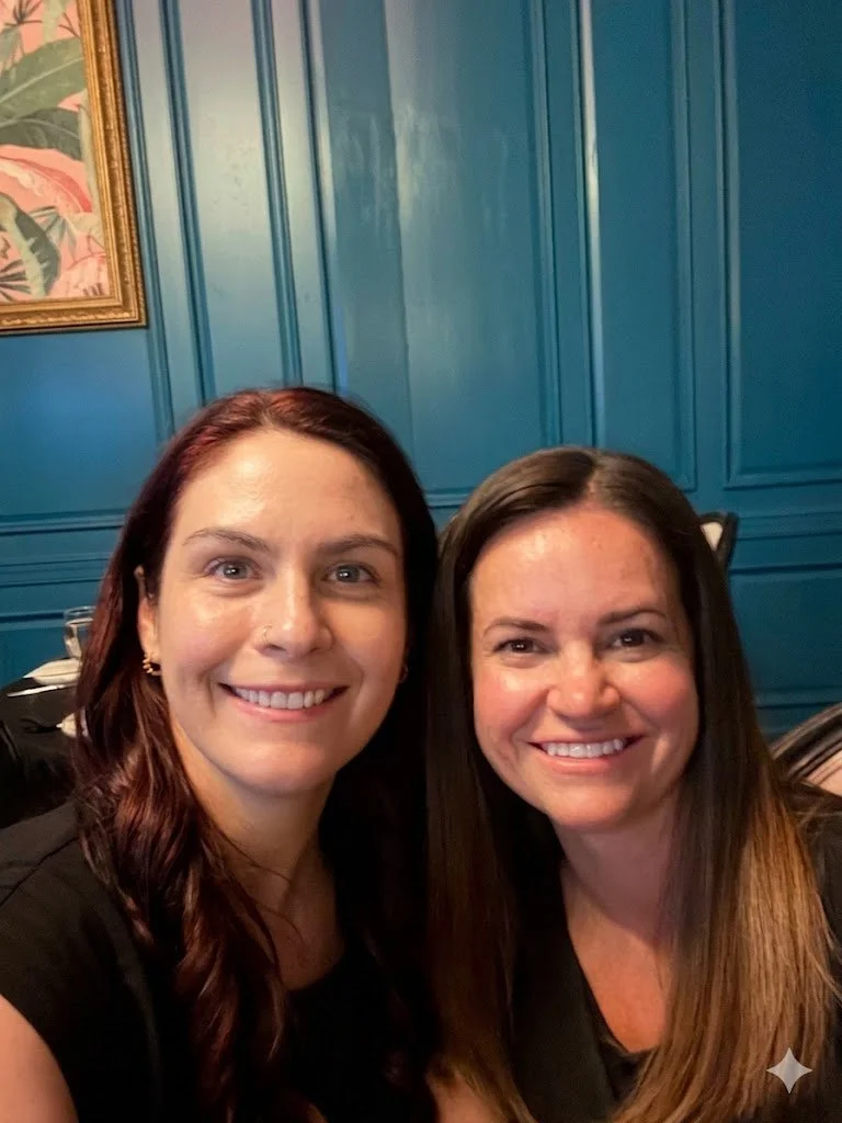 Two women smiling and posing for a photo in a room with blue-paneled walls and a framed botanical picture.