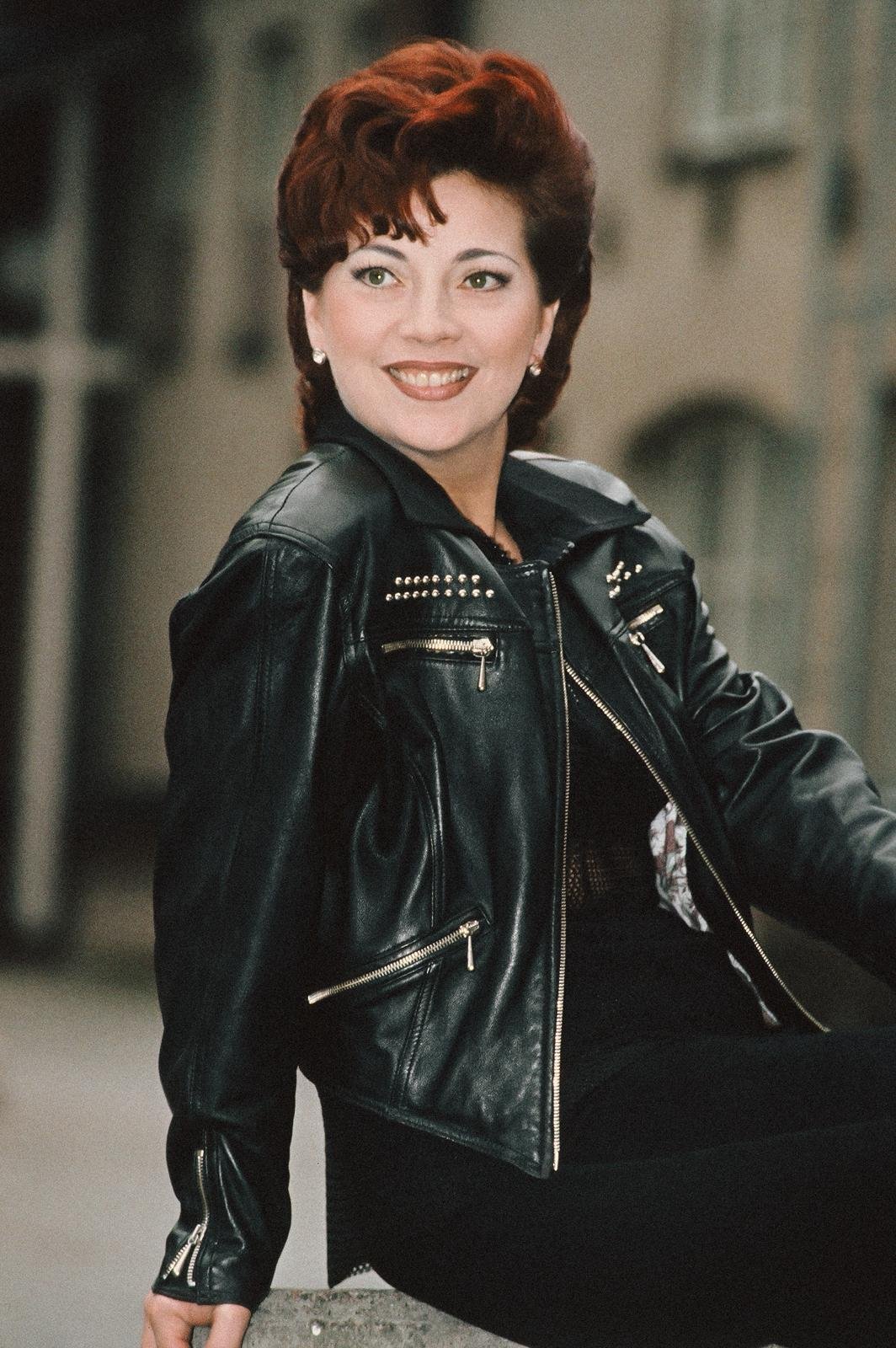 Kristiina Mäki istuu, hymyilee, musta nahkatakki päällä, taustalla rakennuksia.