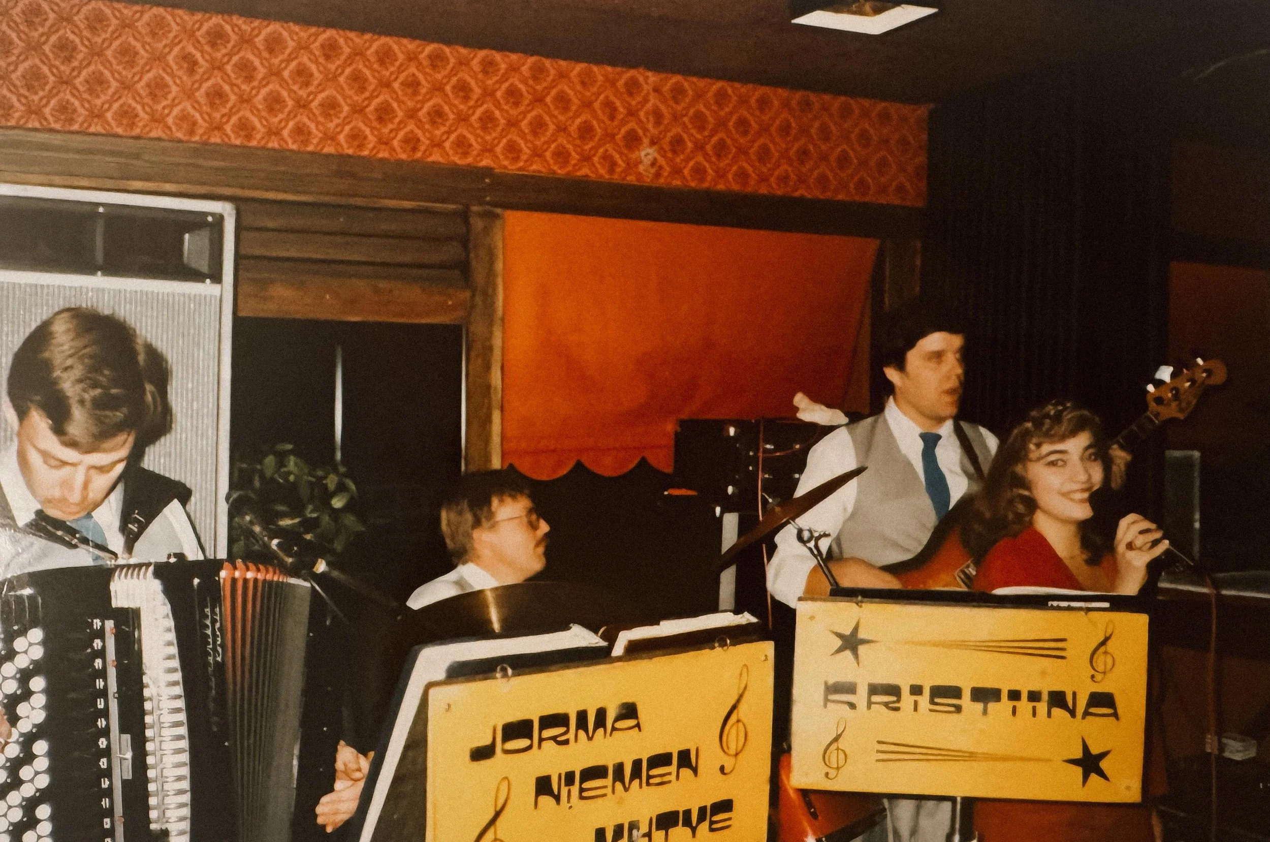 Kristiina Mäki ja Jorma Niemen yhtye soittaa musiikkia, kitaristi, rumpali ja haitaristi esiintyvät, edessä keltaiset kyltit joissa lukee "Jorma Niemen yhtye" ja "Kristiina".