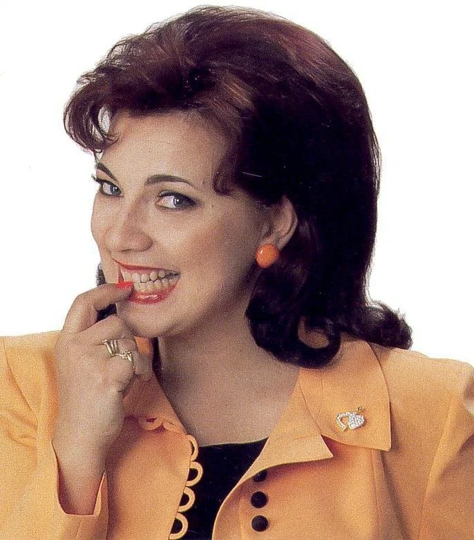 Kristiina Mäki hymyilee ja koskettaa hampaitaan, hänellä on tummanruskeat kiharat hiukset ja oranssi jakku ja korvakorut. Pikkujuttu kuvakortti.