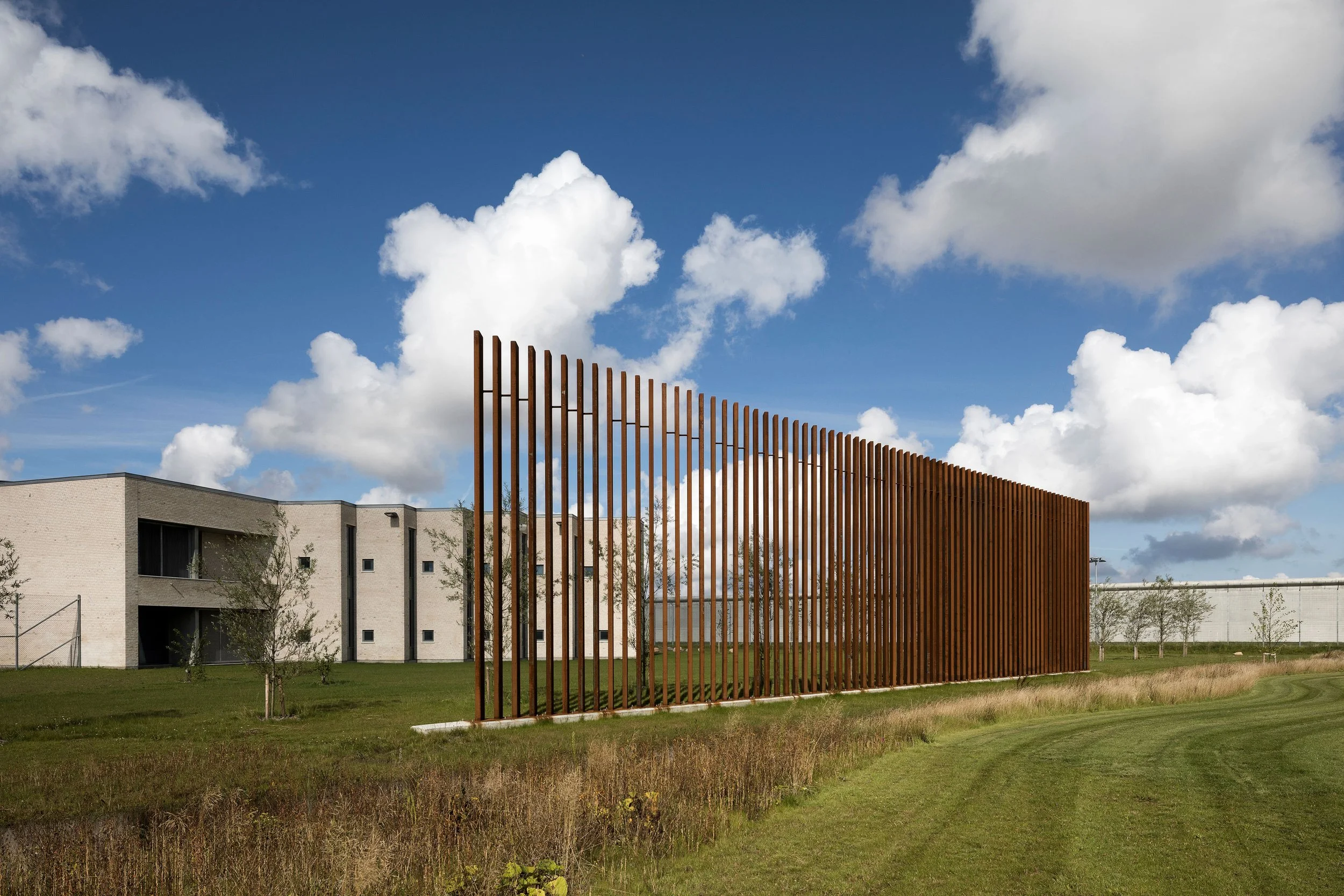 A modern outdoor scene featuring a tall rust-colored vertical metal slat fence in front of a grassy area, with contemporary white residential buildings and a blue sky with scattered clouds in the background.
