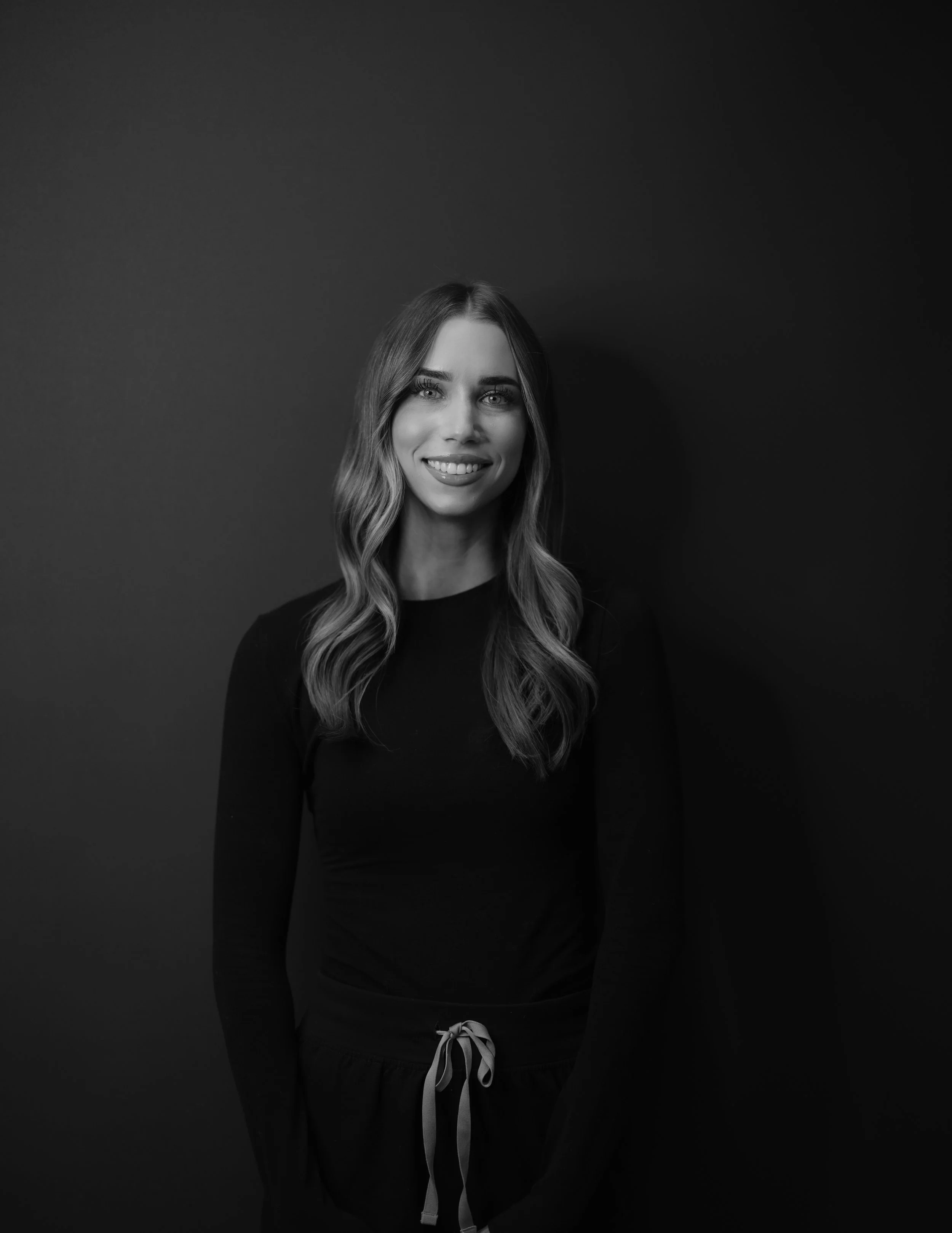Black and white portrait of a young woman smiling, standing against a dark background, wearing a long-sleeved top and drawstring pants.