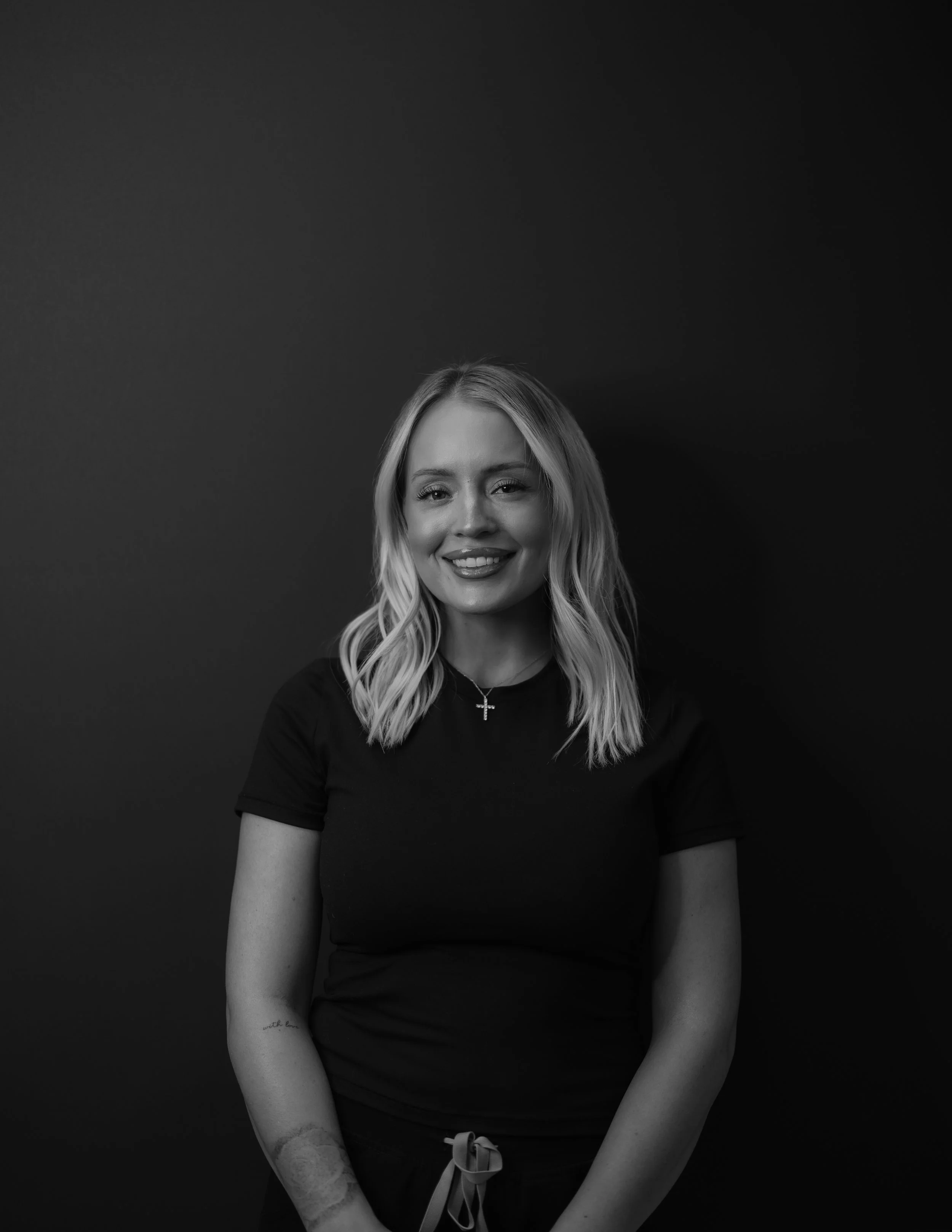 Black and white portrait of the owner of Mrakki Brow Bar with wavy blonde hair, wearing a black t-shirt and a cross necklace, standing against a dark background.