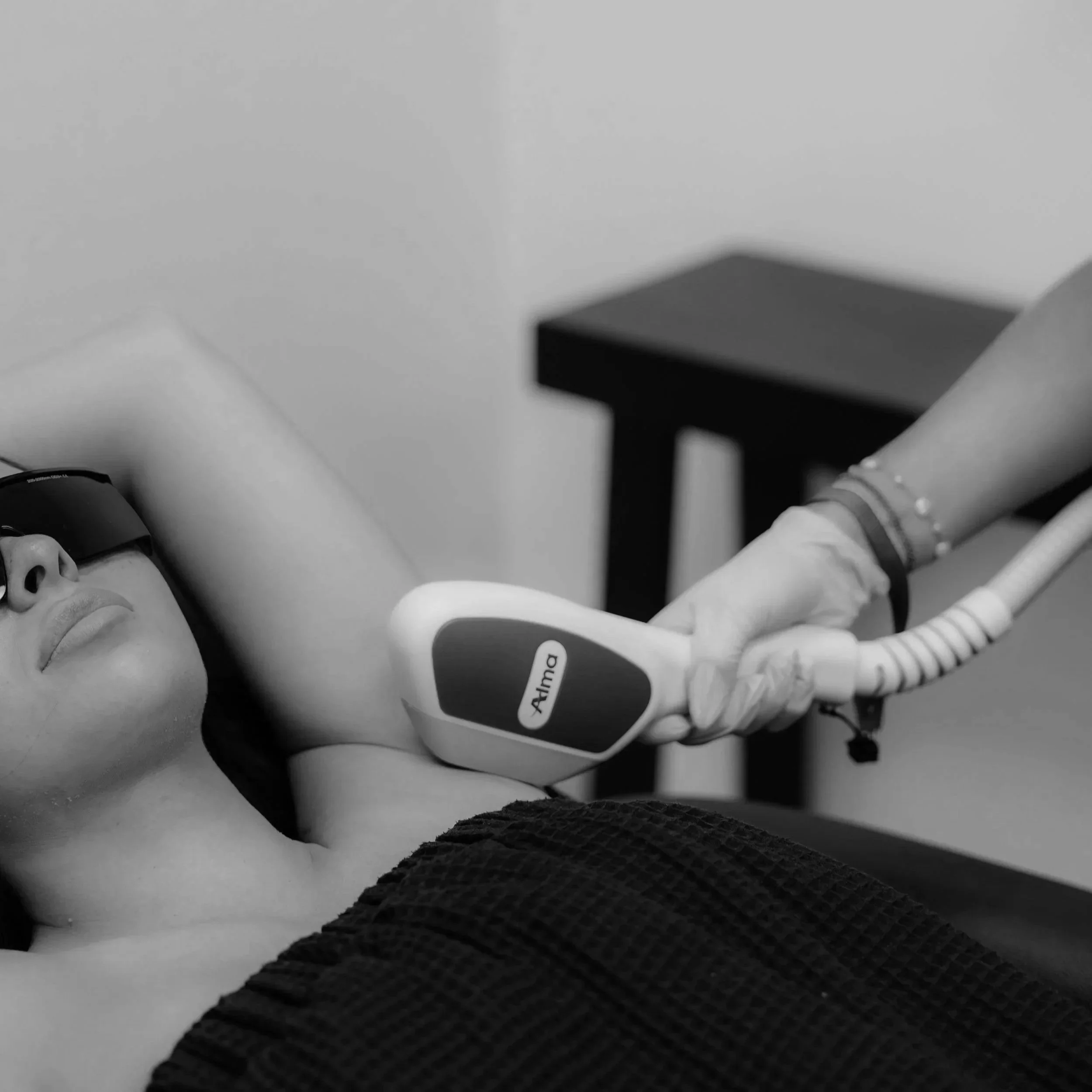 A woman lying on a treatment table wearing eye protection receives a laser treatment on her underarm from a medical professional wearing gloves.