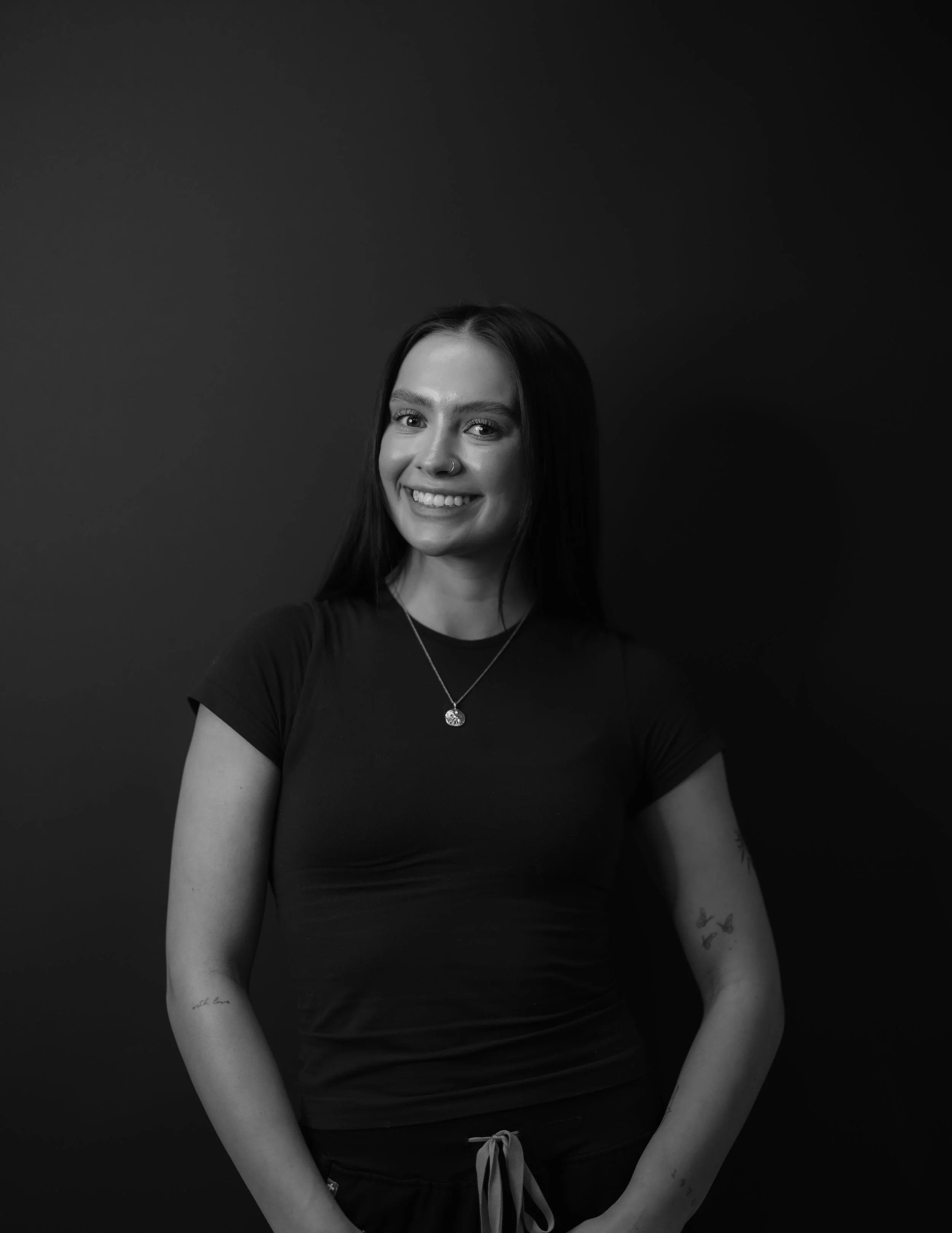 Black and white photo of a smiling young woman with long dark hair, wearing a black t-shirt, a necklace, and tattoos on her arms, standing against a dark background.