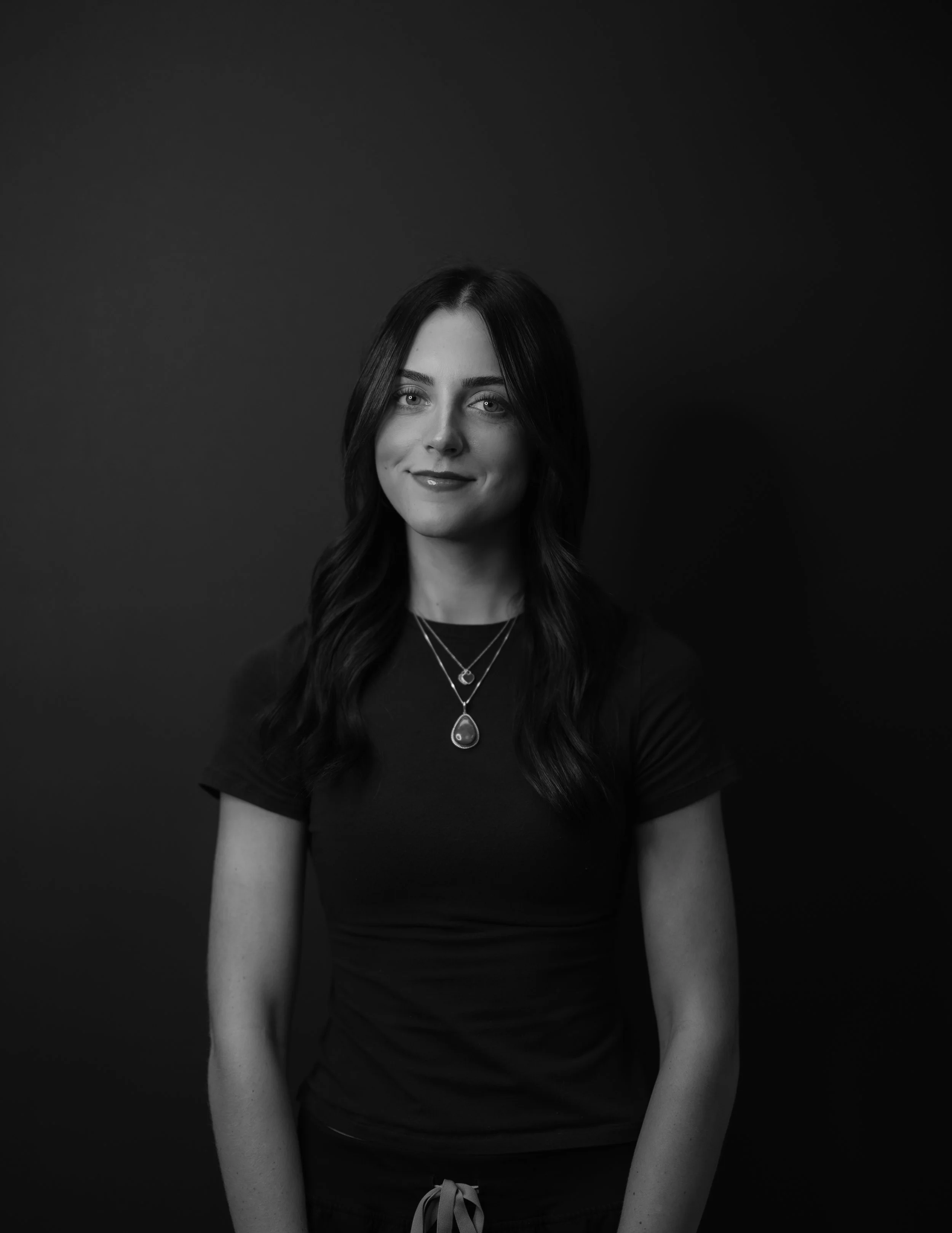 Black and white portrait of a young woman with dark hair, wearing a black t-shirt and layered necklaces, smiling slightly against a dark background.