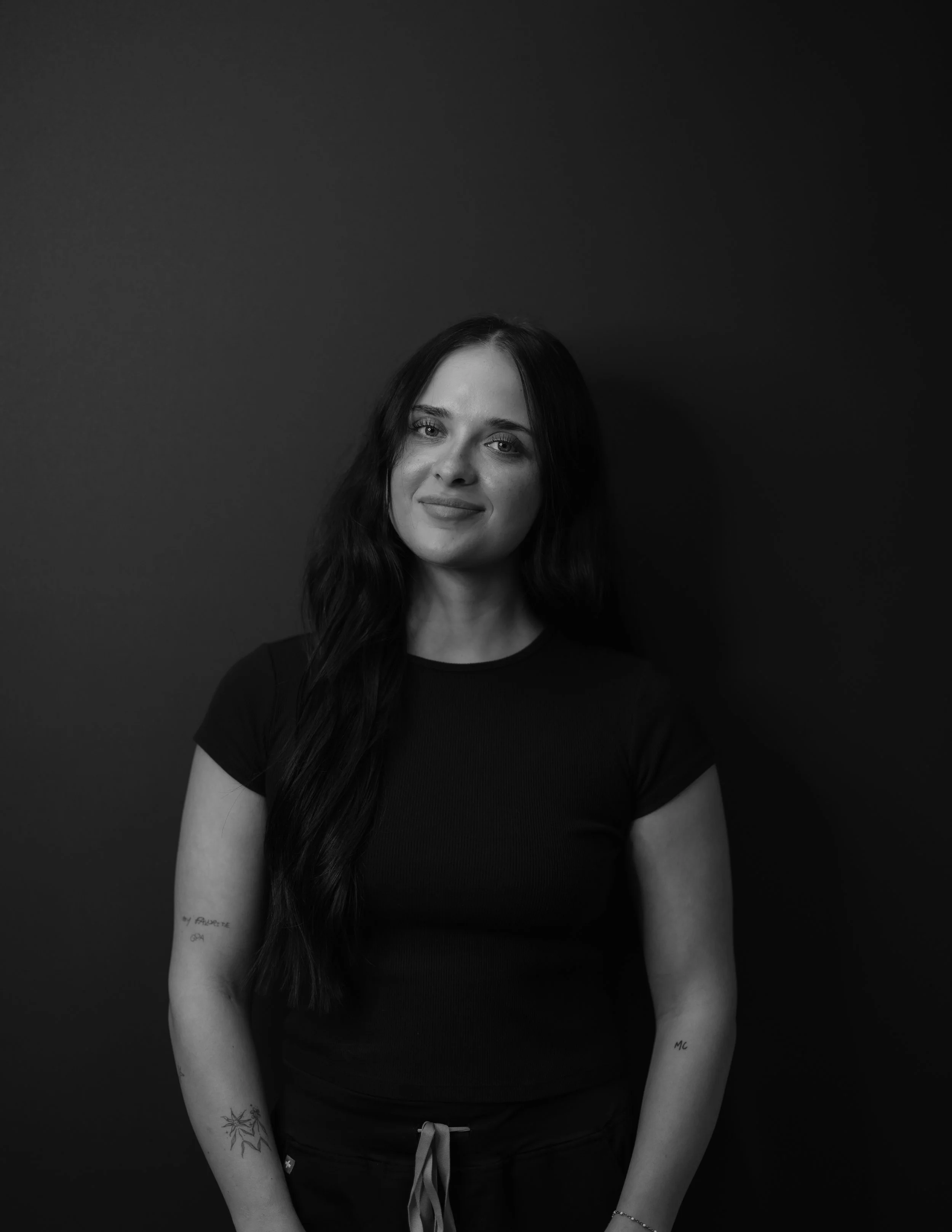Black and white portrait of a woman with long dark hair, wearing a black t-shirt and dark pants, standing against a dark background.