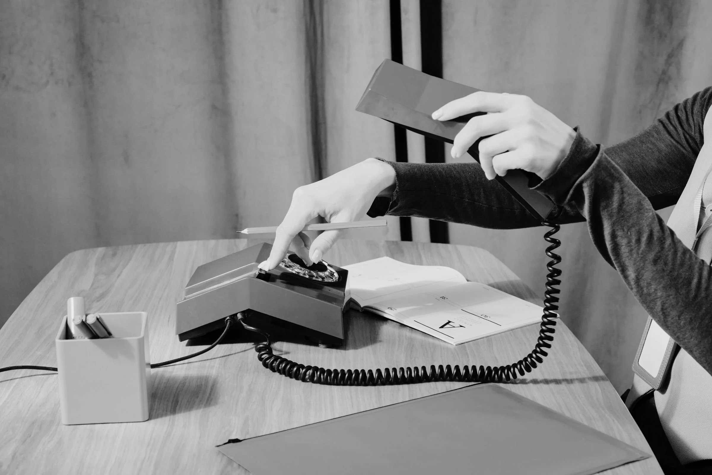 Person with a sleeve on a shirt is holding a phone and a pen at a desk, with a notepad, a small container with pens, and an old-fashioned telephone.