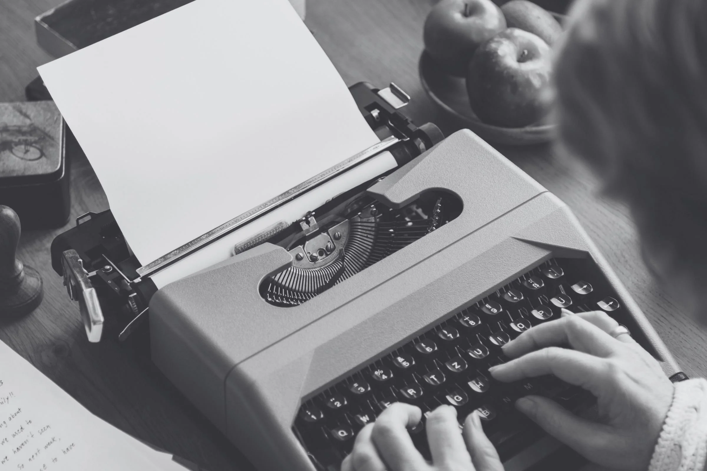 A vintage typewriter with a sheet of paper inserted, being typed on by person with hands visible.