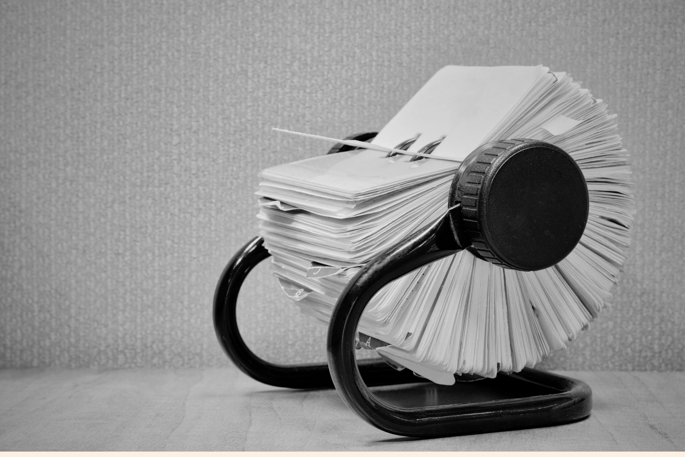 A black and white image of a spiral notebook filled with loose papers, resting on a black, rounded stool. The background is a textured wall.