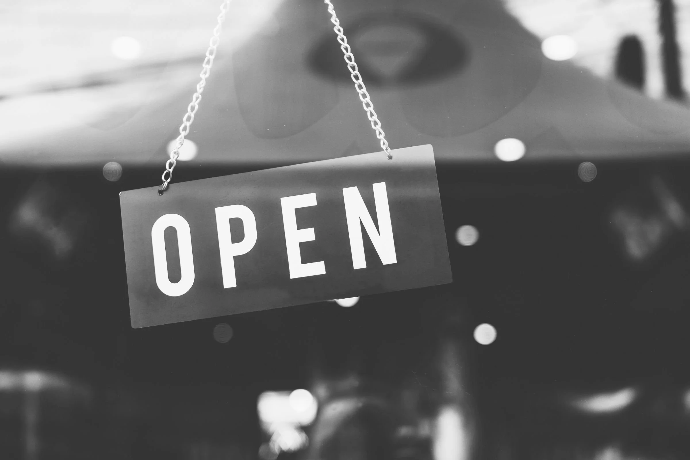 A close-up of a sign hanging on a chain with the word 'OPEN' written in bold white letters, blurred background with bokeh effect.