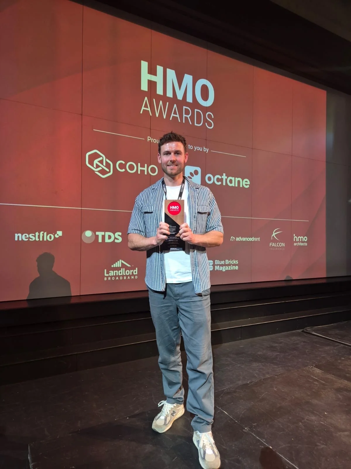 A man holding a trophy at the HMO Awards, standing on stage in front of a large screen displaying the event logo and sponsor names.