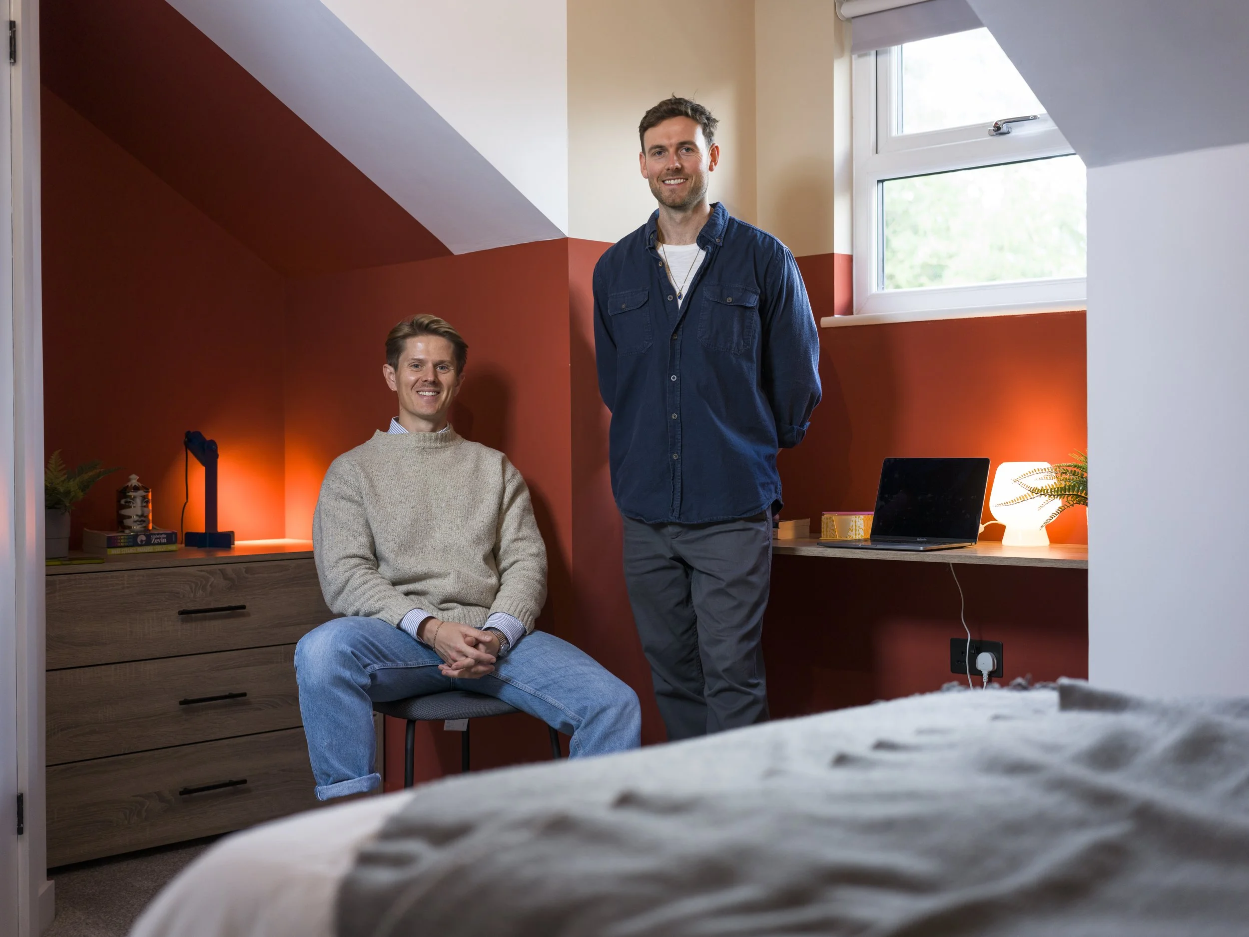 Two young men in a bedroom with a red accent wall, one sitting on a chair wearing a beige sweater and jeans, the other standing in a blue shirt and gray pants, smiling at the camera, with a desk featuring a laptop, a small lamp, and a plant in the background.