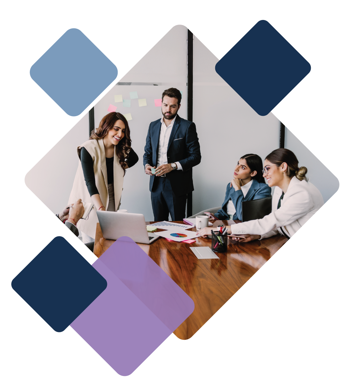 Four colleagues in a business meeting, two women standing and smiling, one woman sitting with glasses and a man standing in a blue suit, all engaged around a table with a laptop, documents, and coffee mugs.