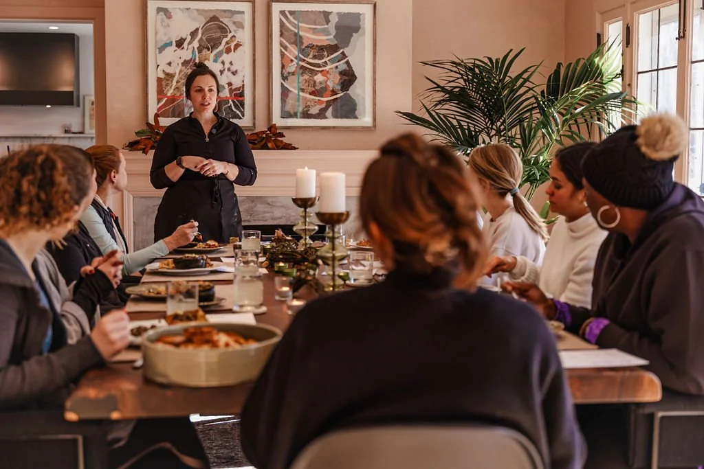 A woman giving a presentation to a group of women seated at a dining table in a cozy, well-decorated room with artwork and large plants.