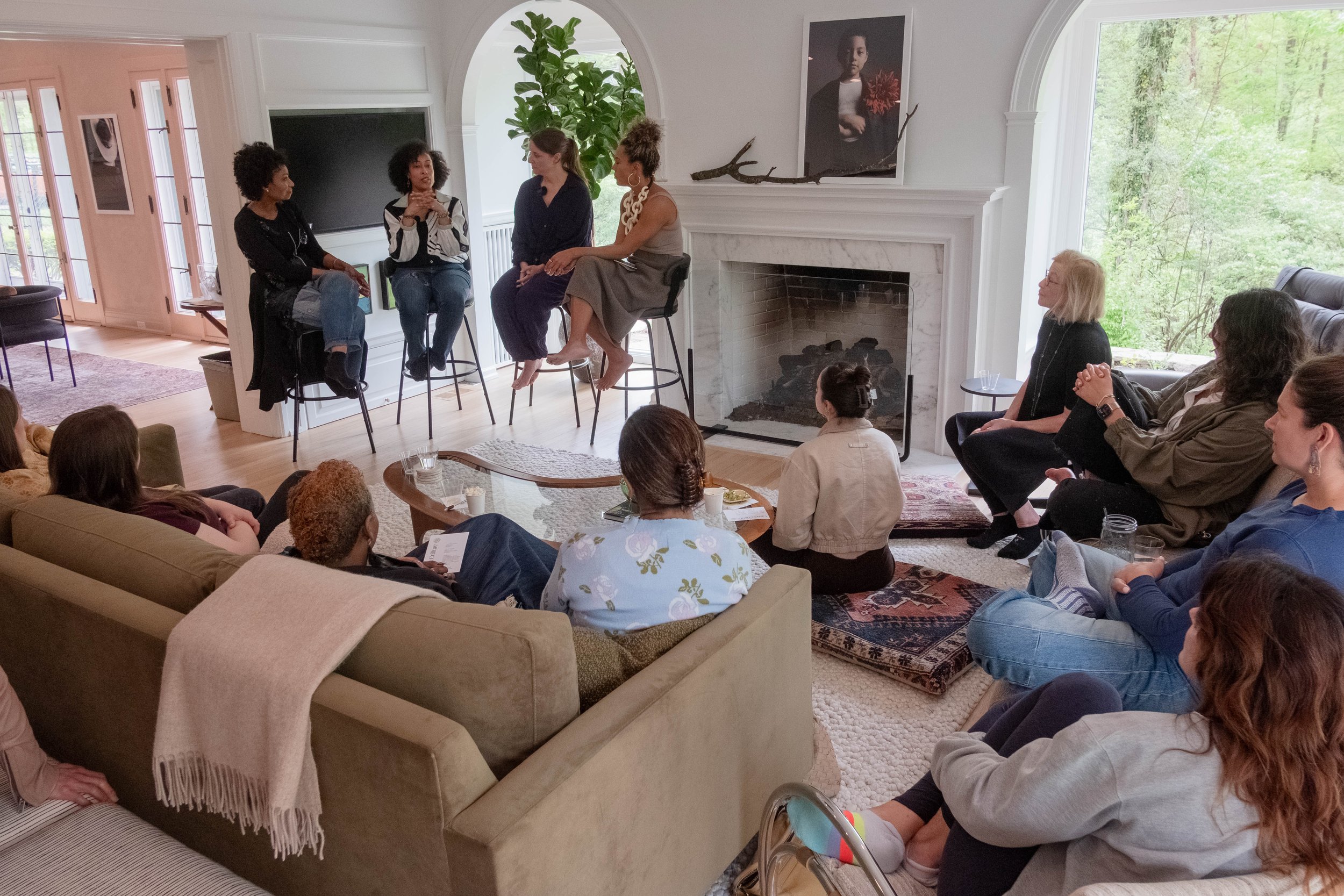 A group of women in a living room participating in a panel discussion. Four women sit on high stools near a fireplace, with an audience seated on sofas and chairs. The room has large windows, a fireplace, and artwork on the walls.