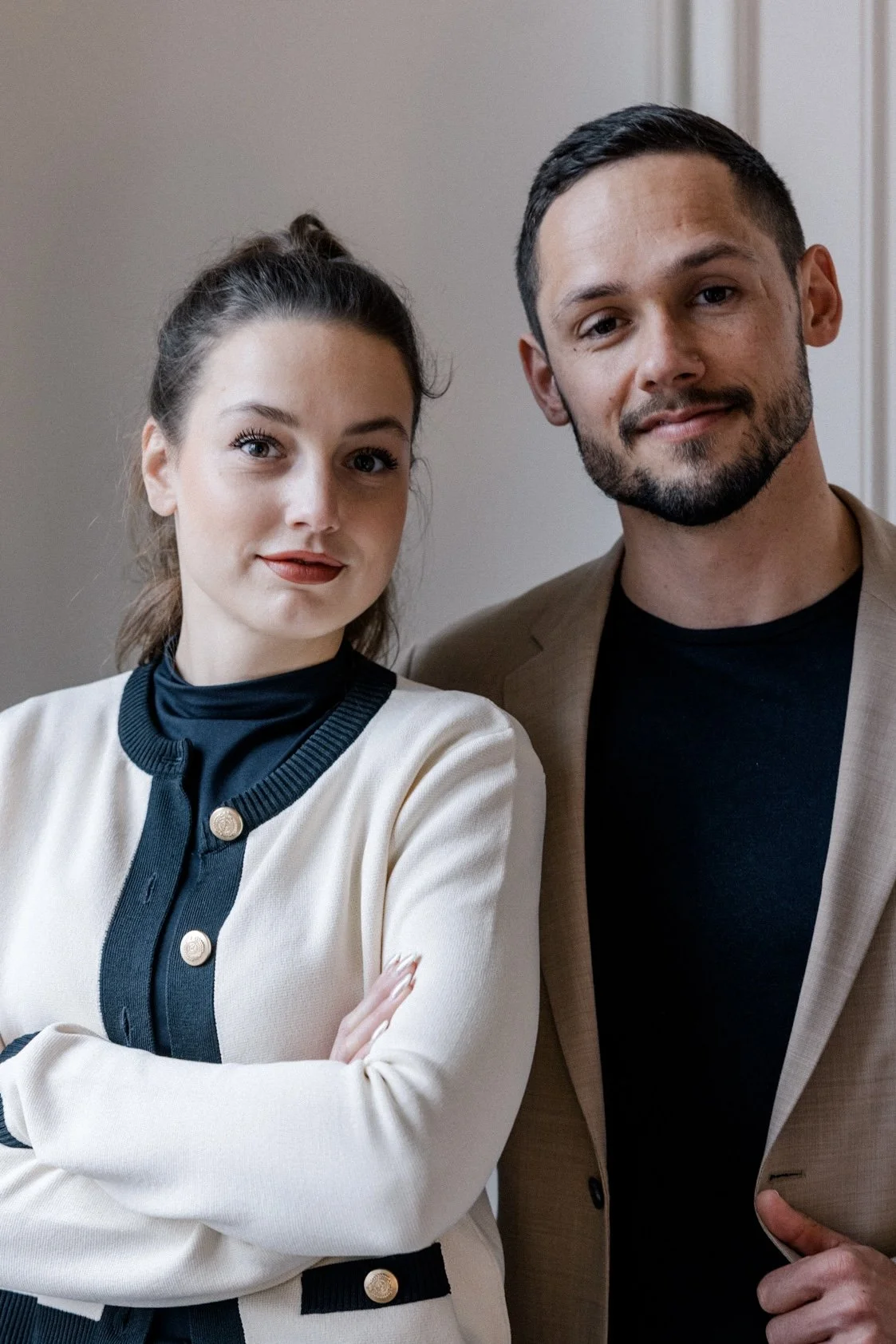 Ein Mann und eine Frau stehen nebeneinander vor einem neutralen Hintergrund, beide schauen in die Kamera und lächeln leicht.