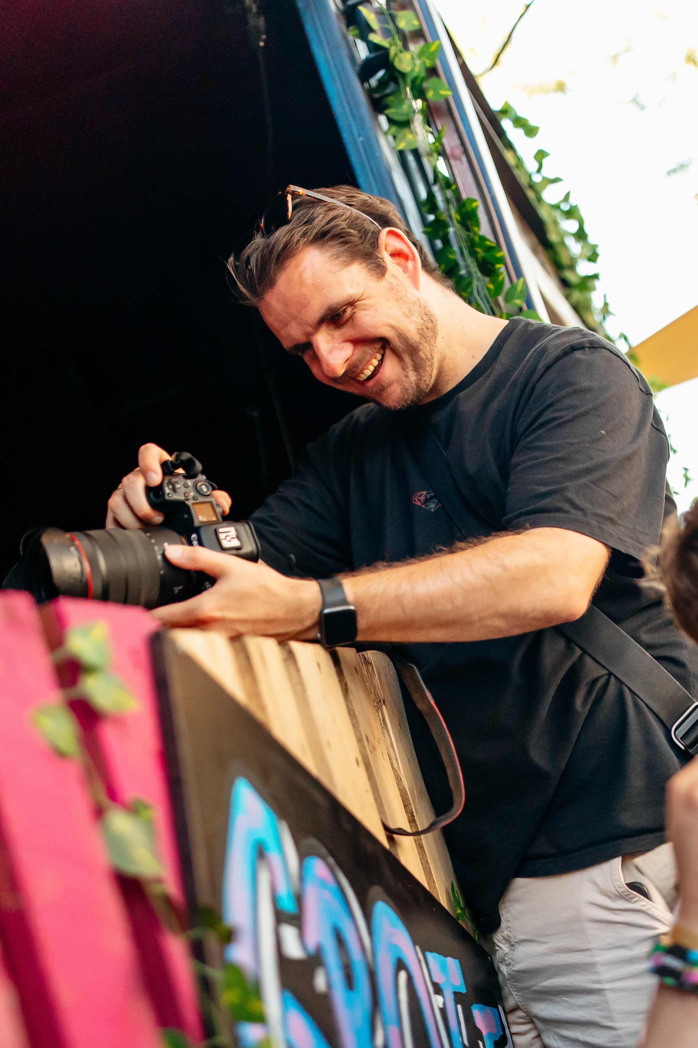Man die lachend met een professionele camera foto's neemt, met een kleurrijk bord en groene bladeren op de achtergrond.