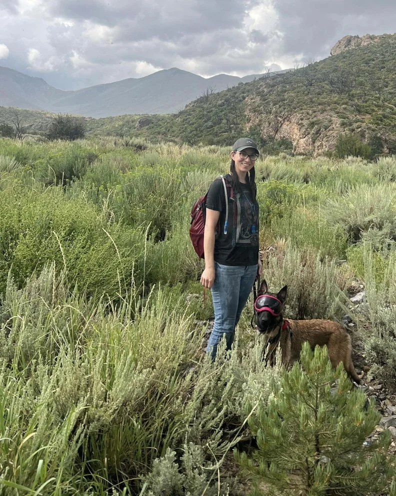 A woman wearing a black t-shirt, jeans, and a cap stands in a grassy field with a brown dog wearing a harness and goggles, with mountains and cloudy sky in the background.
