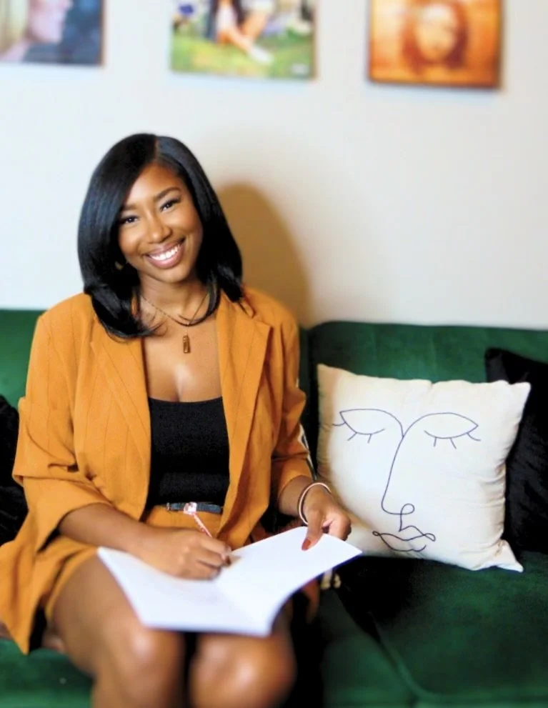 A young woman with black hair, wearing a mustard yellow blazer and black top, sitting on a green couch with a white pillow featuring a minimalist line drawing of a face. She is smiling, holding a notebook, in a room with colorful artwork on the wall.