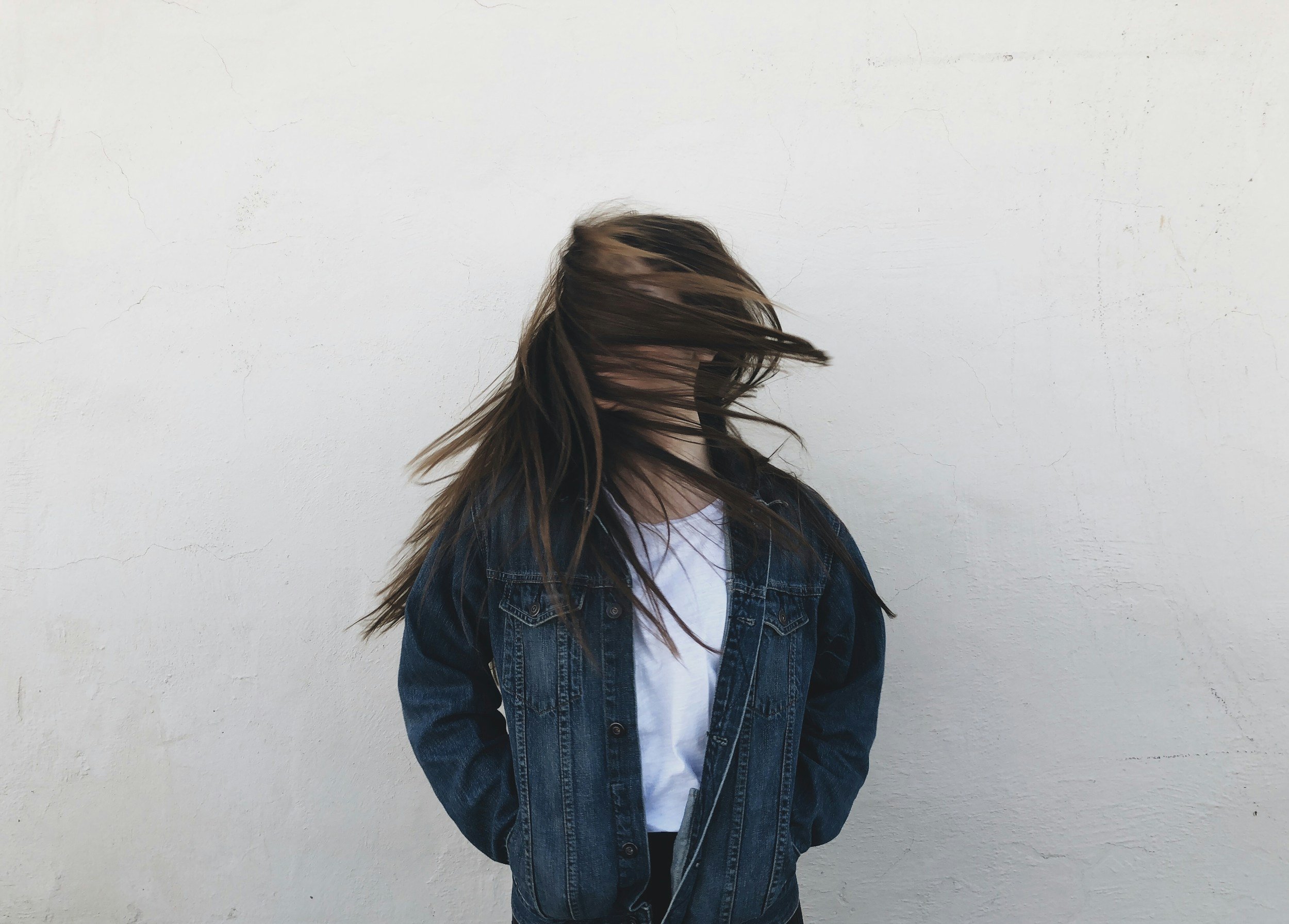Person with long hair moving hair in front of a plain white wall, wearing a denim jacket and a white shirt.