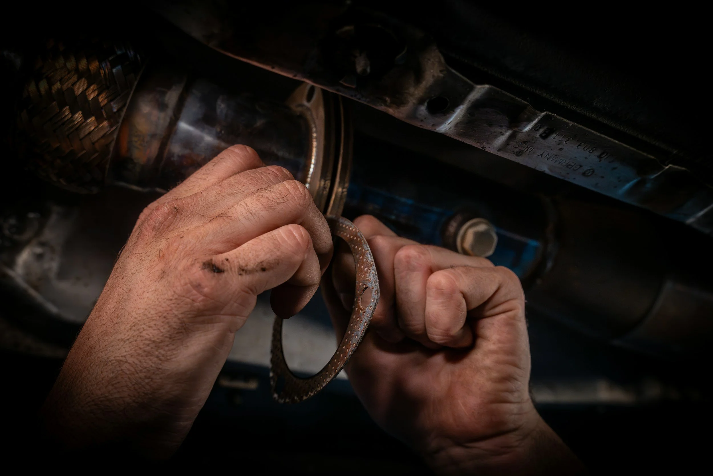 Hands working on a metal component under a vehicle, possibly a mechanic repairing or inspecting car parts.