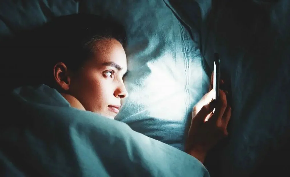 A person lying in bed, looking at a smartphone in a dark room, with the screen illuminating their face.