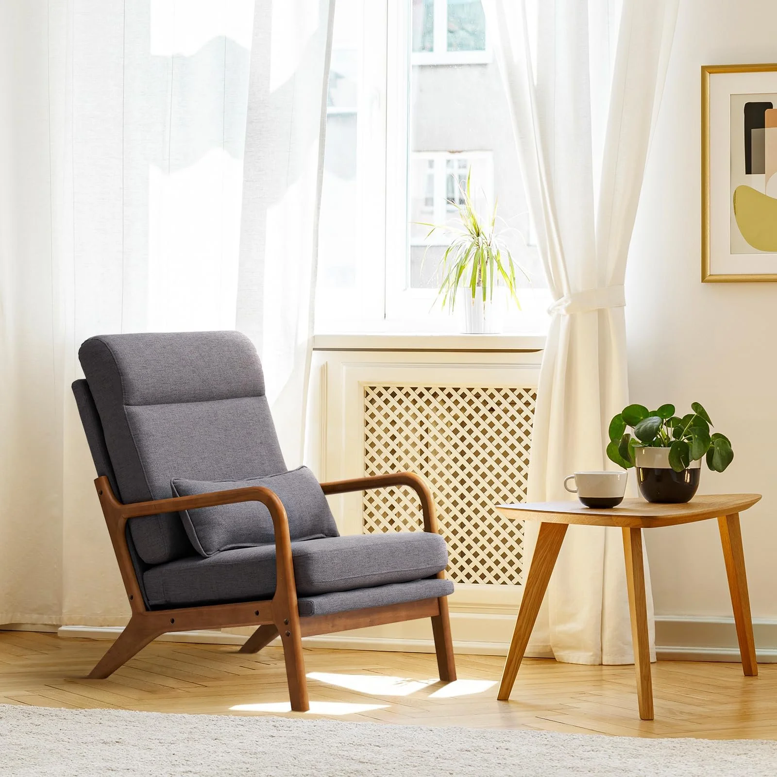 A cozy corner with a gray armchair featuring wooden armrests next to a small wooden table. On the table, there is a black pot with a green plant and a white cup. Behind, a window with white curtains allows natural light to illuminate the room, complemented by framed geometric art on the wall.