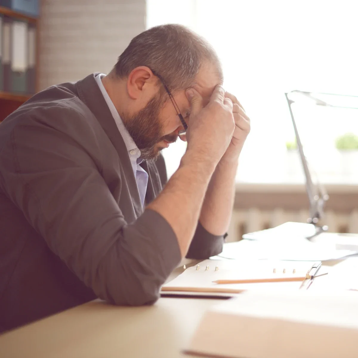 Man with glasses and beard sitting at a desk, holding his head in frustration, surrounded by papers and a pen, with a bright window in the background.