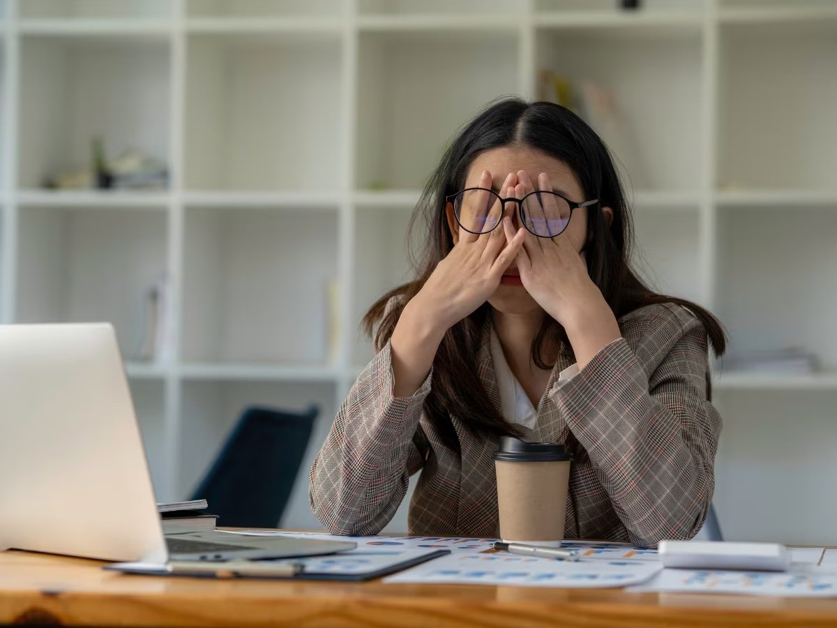 A woman with dark hair and glasses sitting at a desk, holding her face and looking stressed, with a laptop, coffee cup, documents, and office supplies on the desk, in an office setting.