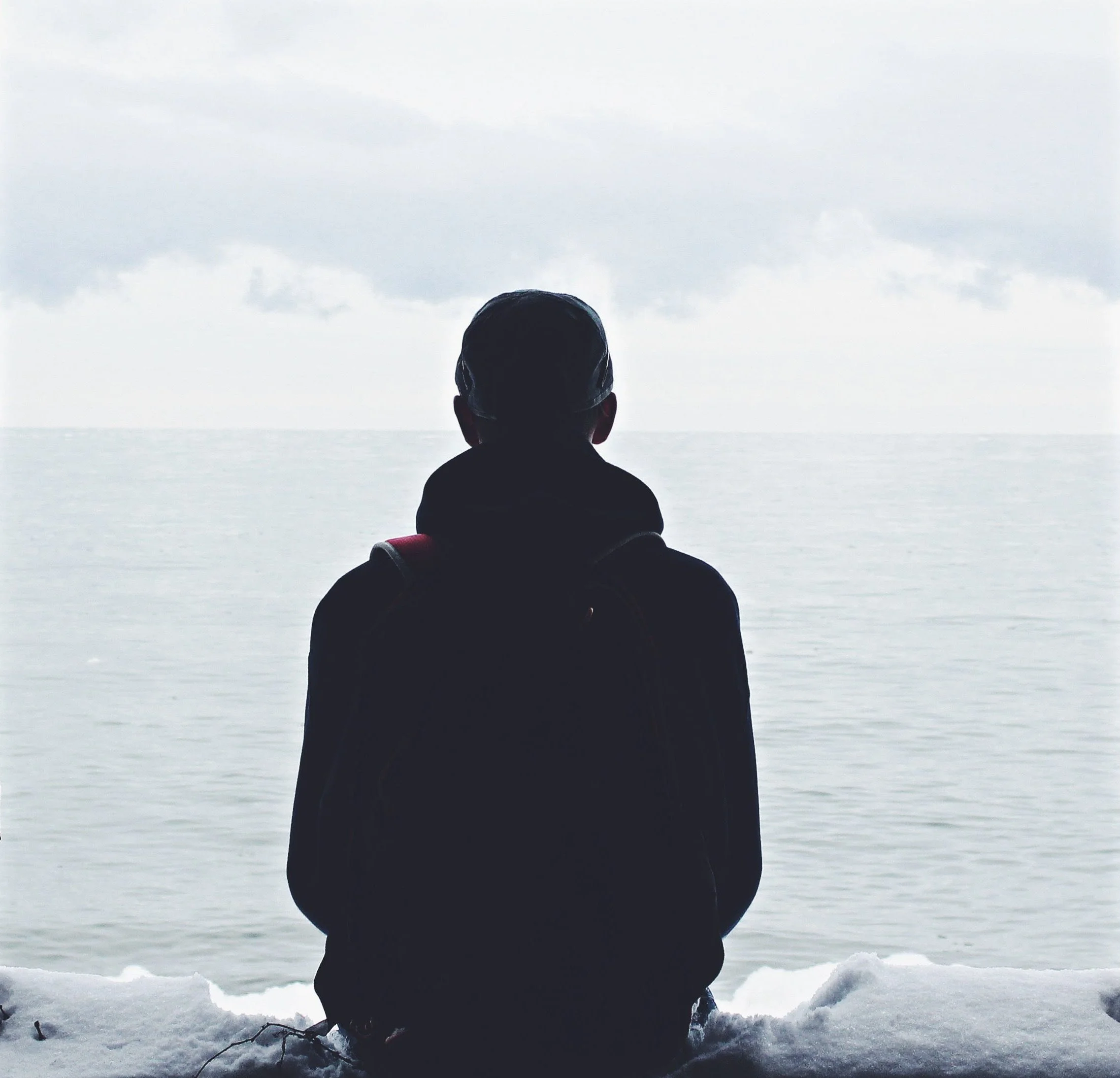 Person sitting on snow while looking at cloudy sky and distant horizon over water.