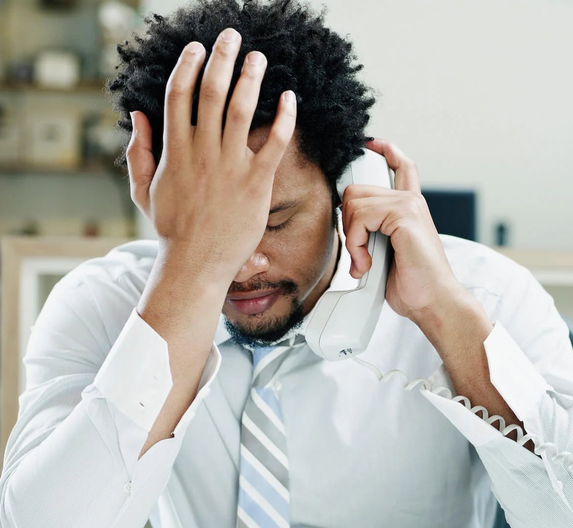 Man in white shirt and tie holding his head in stress while talking on landline phone in an office.