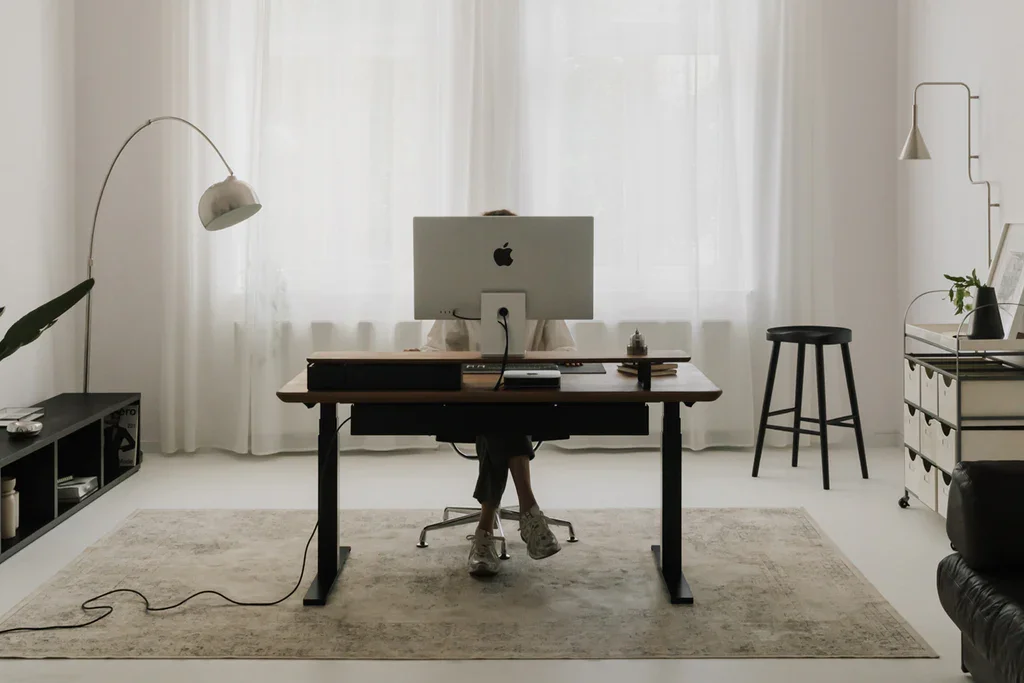 Minimalist home office with a person working at a desk, an iMac computer, a standing lamp, and decorative plants in a bright room with white curtains.